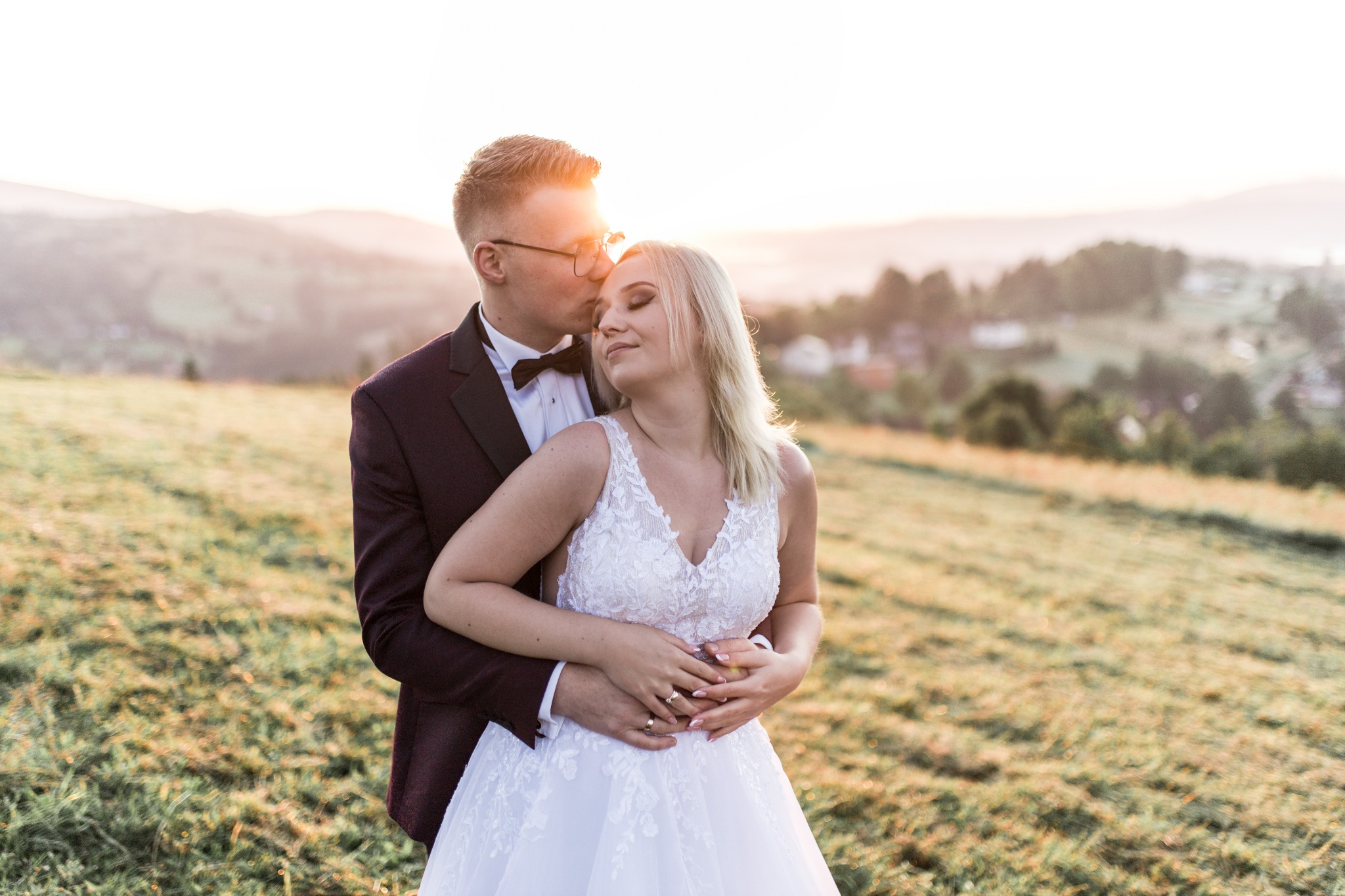Para młoda w objęciach na tle wschodzącego słońca i pagórkowatego krajobrazu; mężczyzna w bordowym smokingu całuje kobietę w białą suknię ślubną.