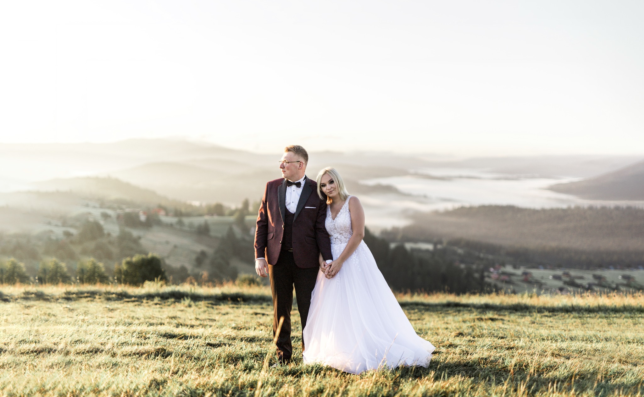 Para młoda w strojach ślubnych stoi na łące z widokiem na góry we mgle o wschodzie słońca, panna młoda opiera głowę o ramię pana młodego.