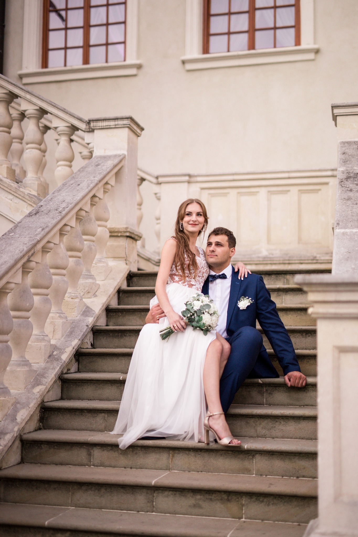 Uśmiechnięta para w strojach ślubnych siedzi na kamiennych schodach pałacu, panna młoda trzyma bukiet.