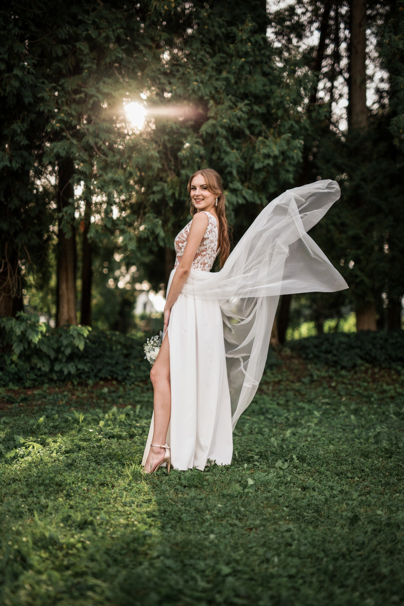 Uśmiechnięta panna młoda w koronkowej sukni z rozcięciem, pozuje w plenerze z powiewającym welonem, trzymając bukiet polnych kwiatów.