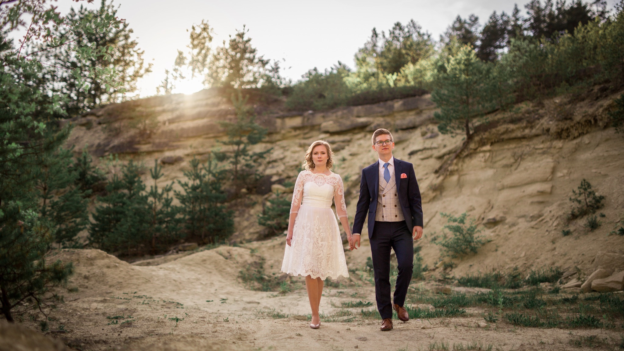 Para młoda trzymająca się za ręce spaceruje po piaszczystym terenie z drzewami w tle, mężczyzna w granatowym garniturze z kamizelką w kratę, kobieta w białej koronkowej sukience do kolan.