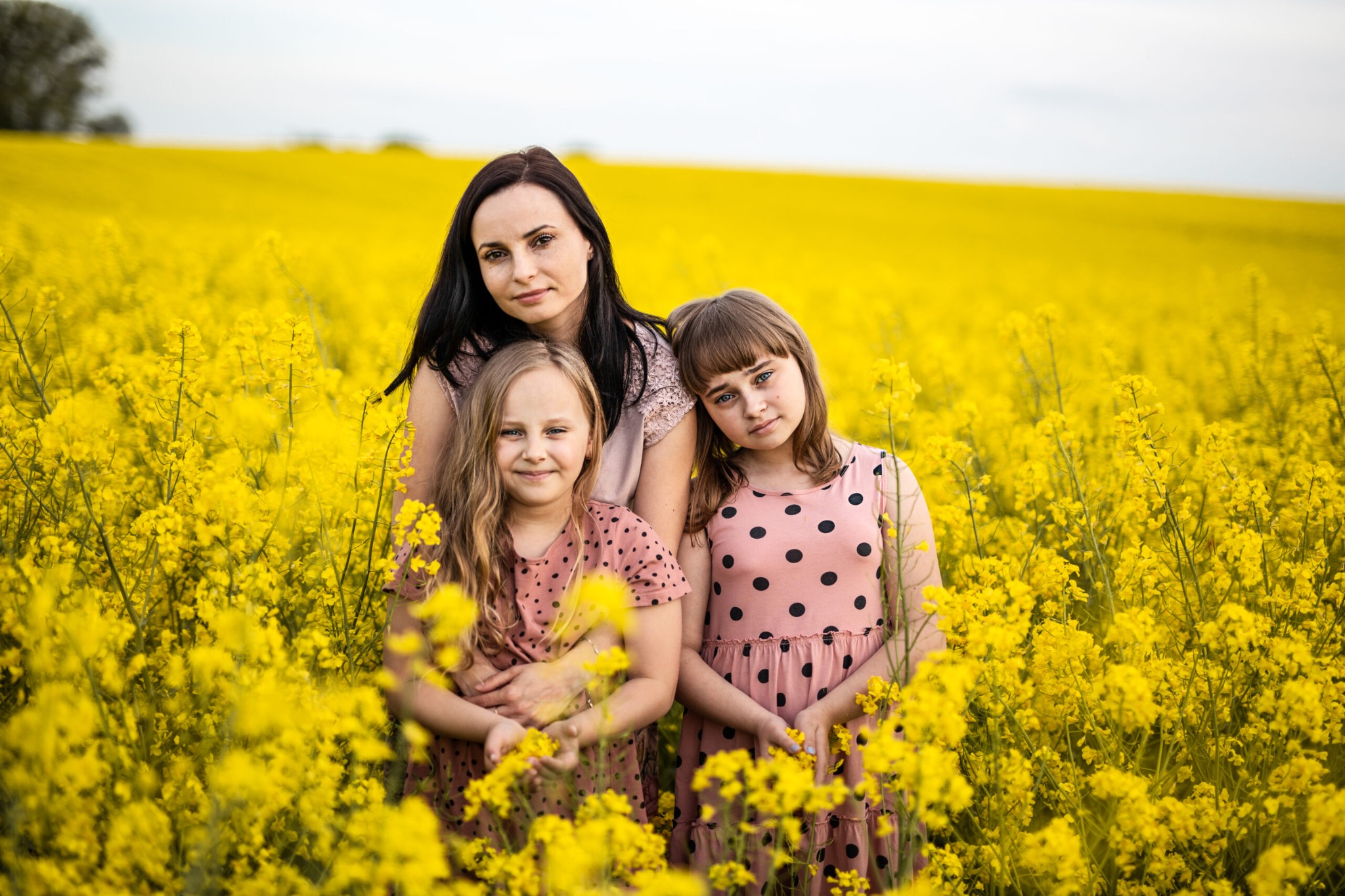 Portret matki z dwiema córkami w kwitnącym żółtym polu rzepaku, dziewczynki w różowych sukienkach w czarne kropki, stonowana kolorystyka.