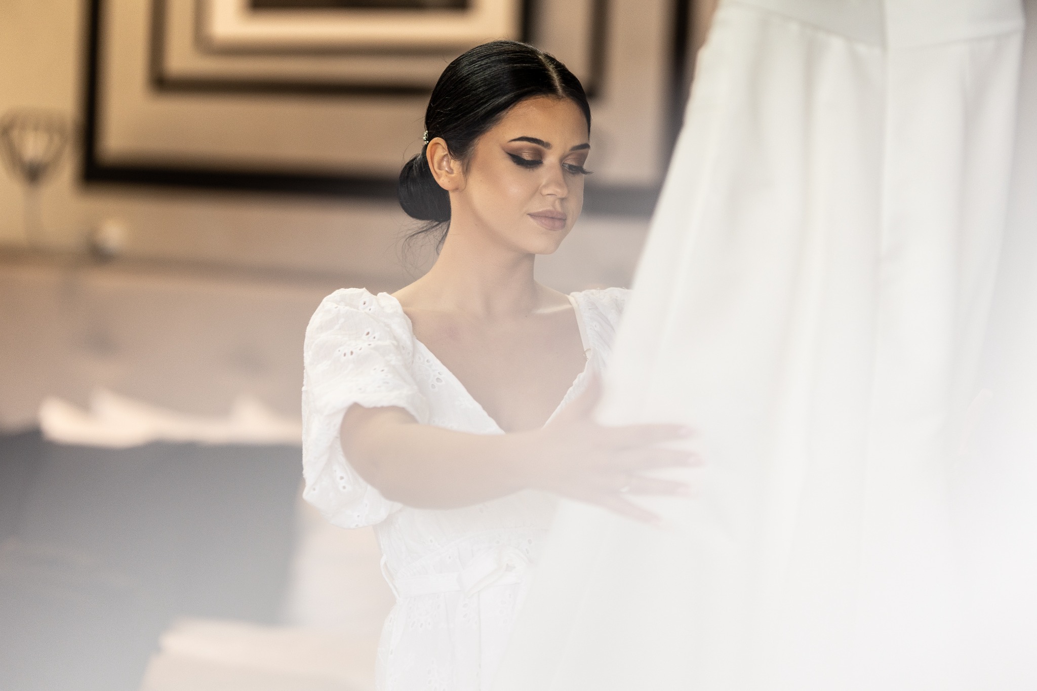 Elegancka kobieta w białej sukience, z upiętymi ciemnymi włosami i subtelnym makijażem, delikatnie dotyka białej sukni ślubnej w jasnym wnętrzu.