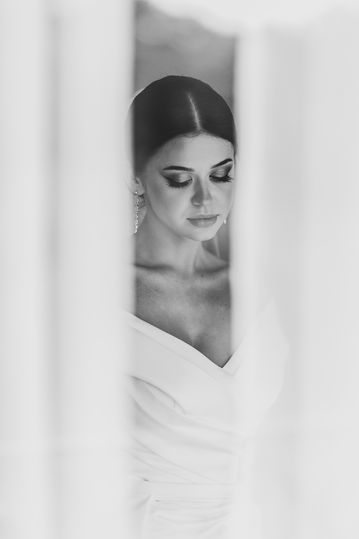 Czarno-białe zbliżenie panny młodej z zamkniętymi oczami, widoczna przez delikatne, rozmyte firanki, eksponujące makijaż i elegancką suknię.