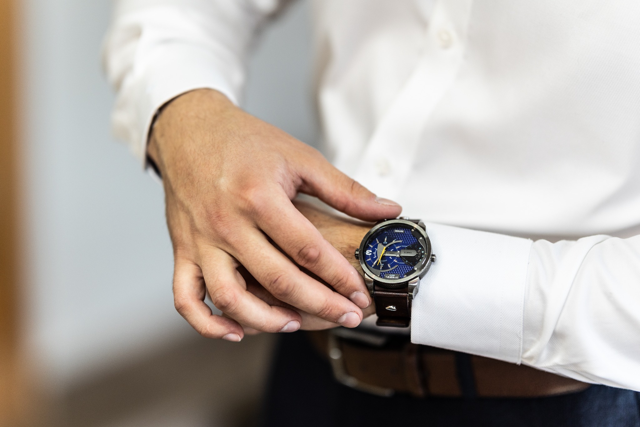 Męska dłoń z zegarkiem Diesel na skórzanym pasku, biała koszula z mankietami, brązowy pasek od spodni.