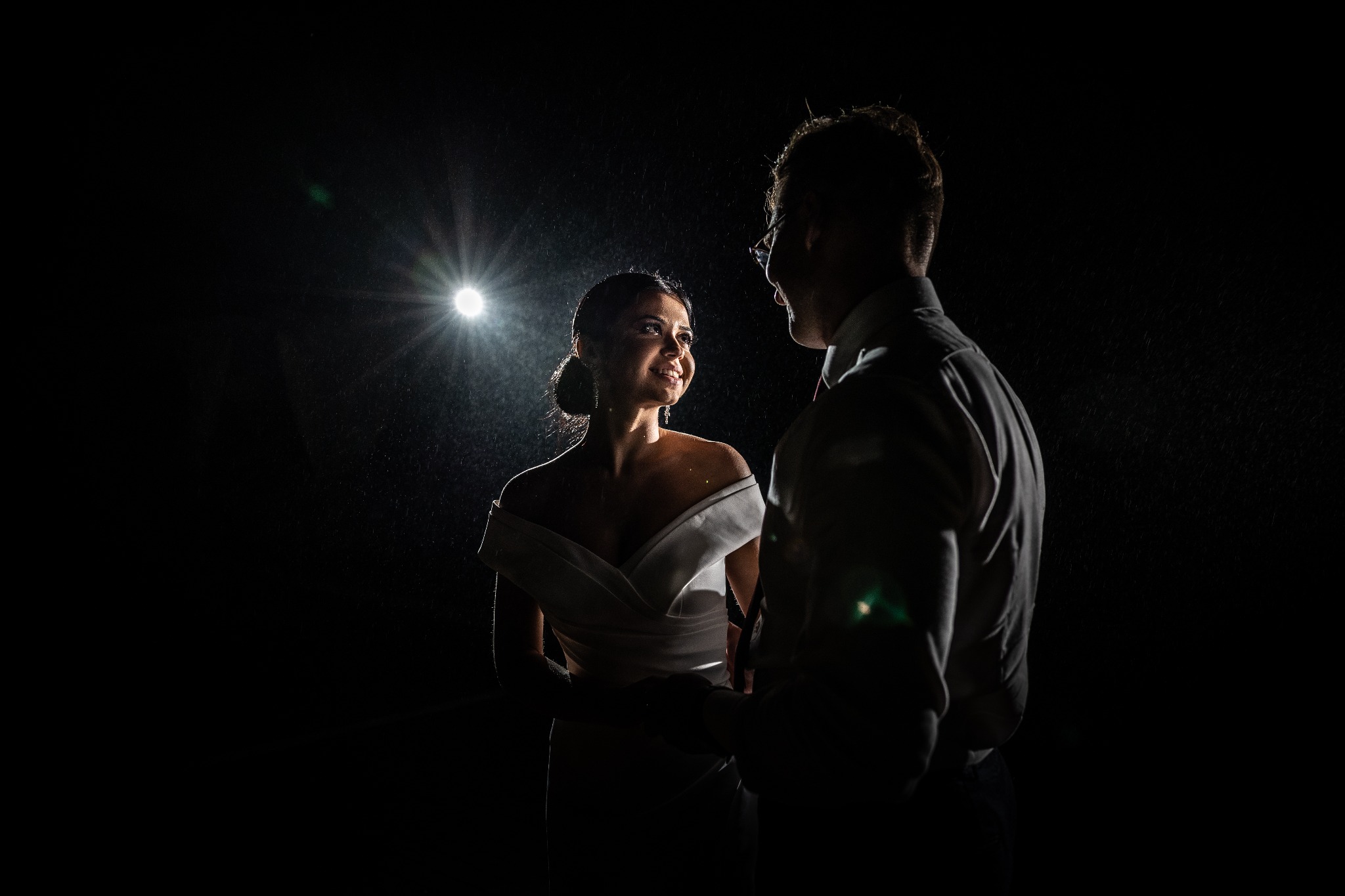 Para młoda w deszczu, panna młoda w białej sukni z uśmiechem spogląda na pana młodego w okularach, oświetleni od tyłu pojedynczym światłem.