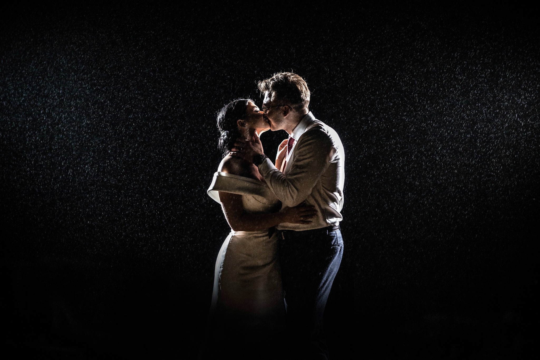 Para całująca się w strugach deszczu na ciemnym tle, panna młoda w białej sukni z odkrytymi ramionami, pan młody w białej koszuli i ciemnych spodniach.