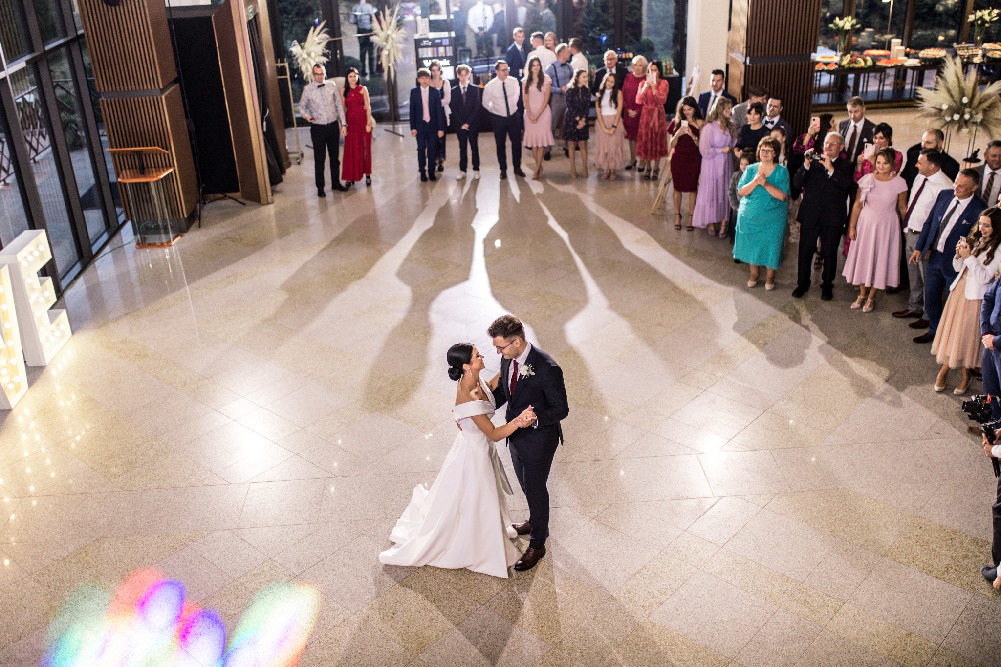 Para młoda tańczy na parkiecie podczas wesela, widzowie obserwują, duże litery z żarówkami w tle.