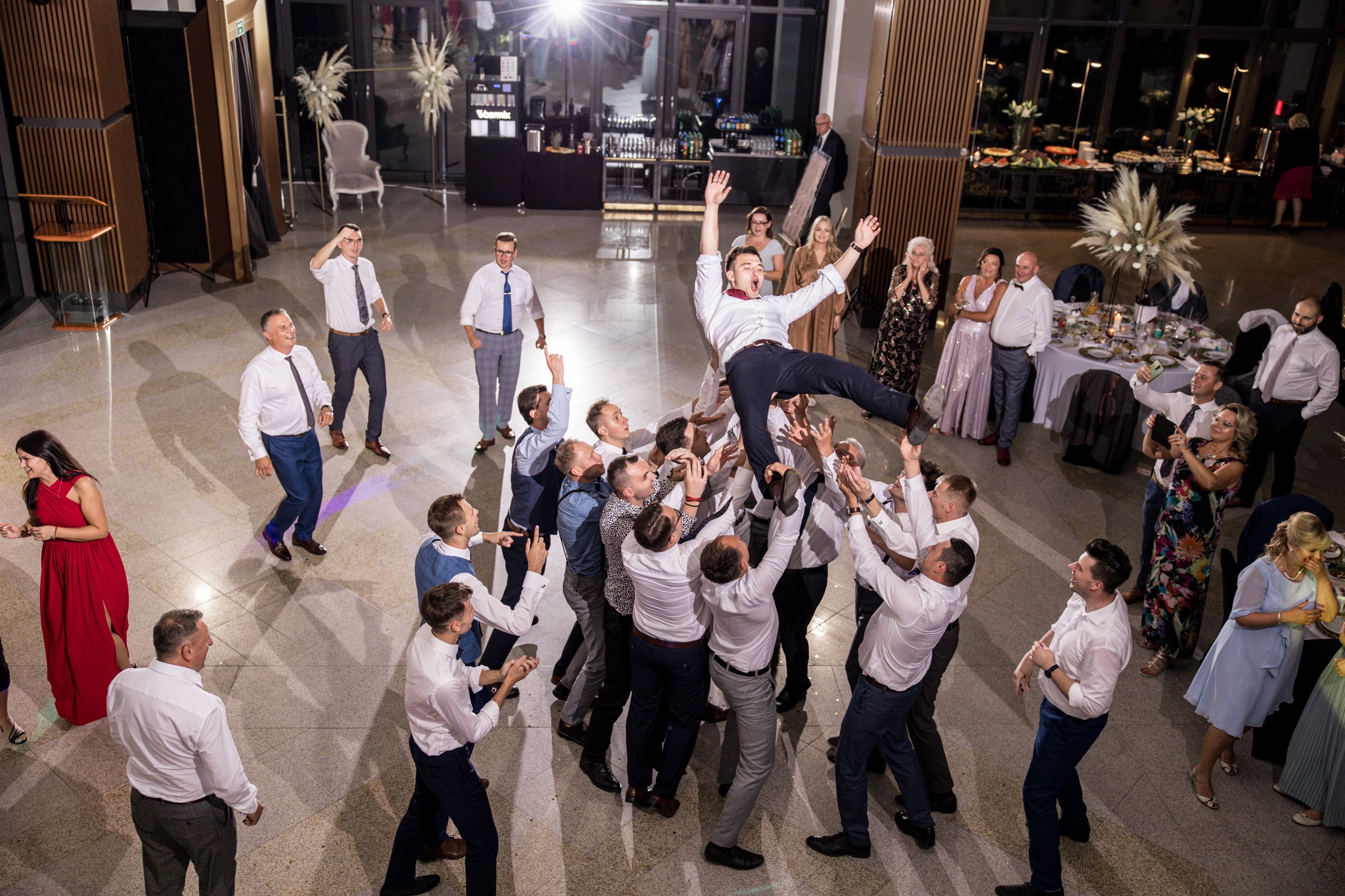 Radosna scena na weselu: grupa mężczyzn podrzuca pana młodego do góry, w tle goście weselni i dekoracje sali.