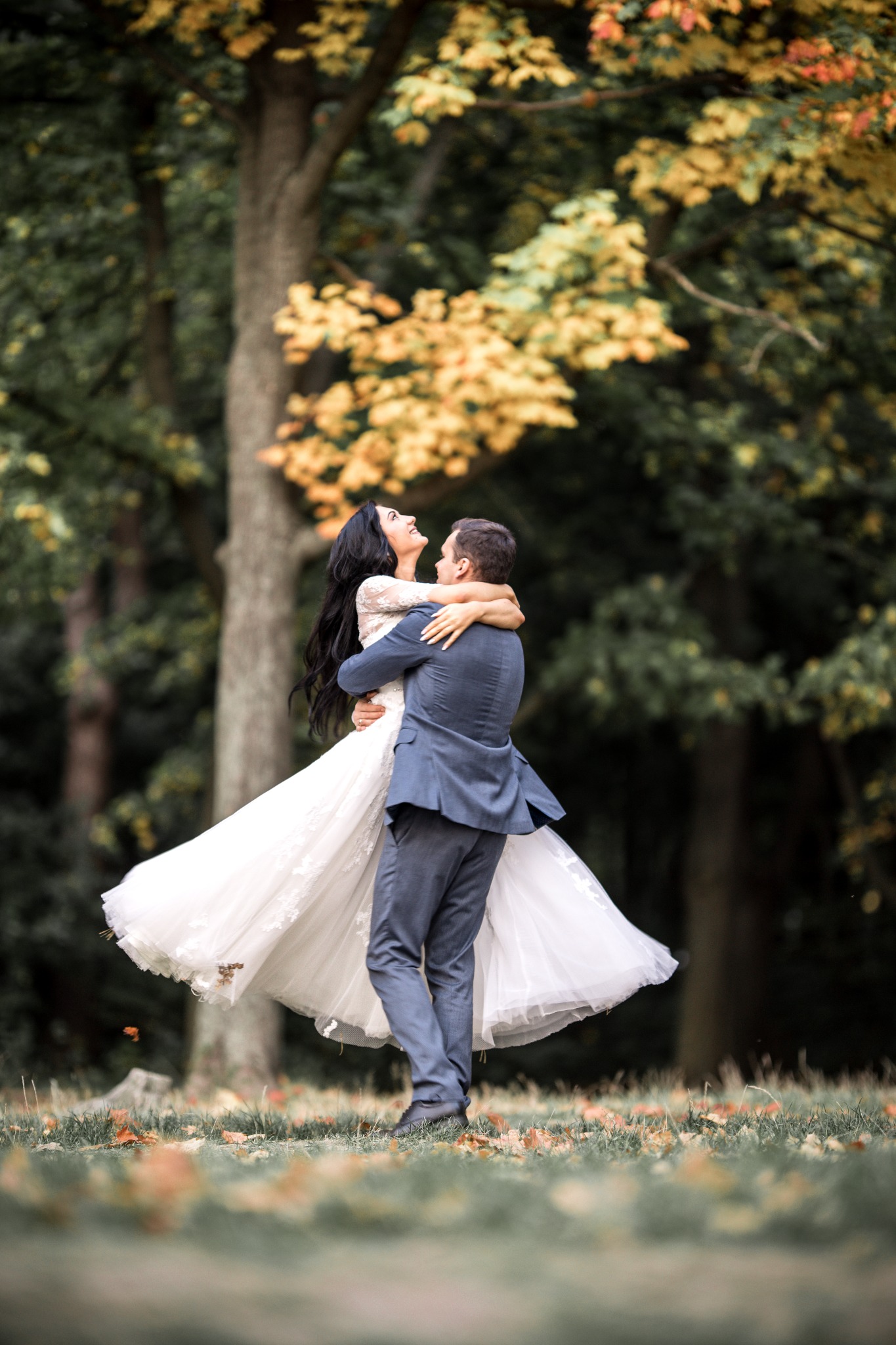 Para młoda w objęciach na tle jesiennego parku z drzewami o żółtych liściach, panna młoda w koronkowej sukni, a pan młody w garniturze.