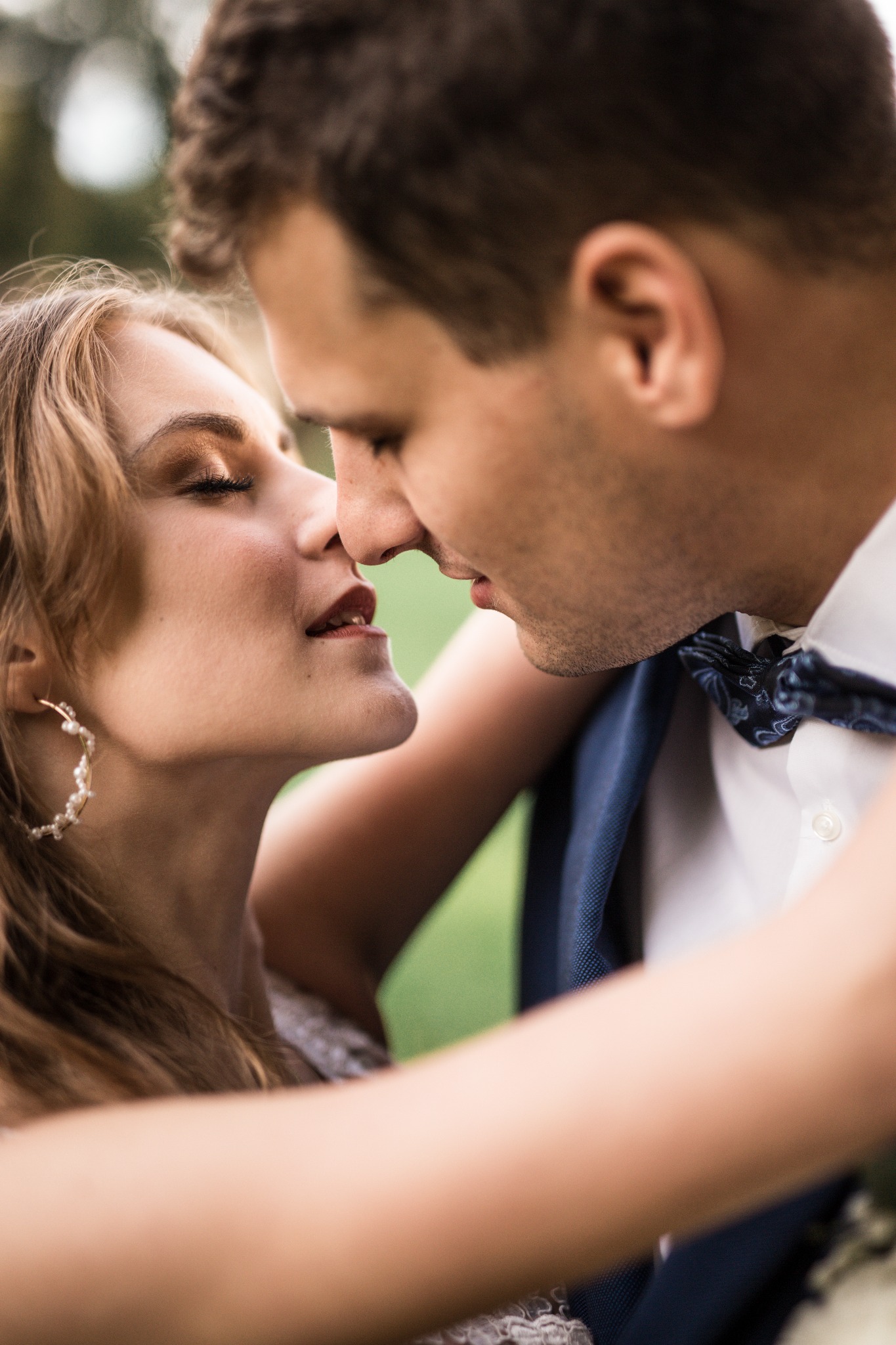 Zbliżenie na parę młodych ludzi, delikatnie dotykających się nosami, panna młoda z zamkniętymi oczami i kolczykami w kształcie półksiężyca z perłami, pan młody w białej koszuli i granatowej muszce...