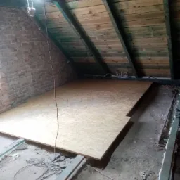Płyta OSB ułożona na poddaszu z ceglaną ścianą i drewnianą więźbą dachową, widoczne kable i prowizoryczne oświetlenie.