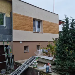 Fragment elewacji domu jednorodzinnego w trakcie prac wykończeniowych z widoczną warstwą izolacji, tynkiem strukturalnym imitującym drewno oraz rusztowaniem i materiałami budowlanymi na pierwszym...