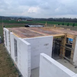 Wznoszenie ścian z białych bloczków na budowie domu jednorodzinnego, widoczne drewniane szalunki i przygotowane deskowanie stropu, w tle krajobraz wiejski.