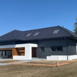 Nowoczesny dom jednorodzinny z czarną elewacją i białymi akcentami architektonicznymi, widoczne okna dachowe i fragment terenu przed budynkiem w trakcie zagospodarowywania.