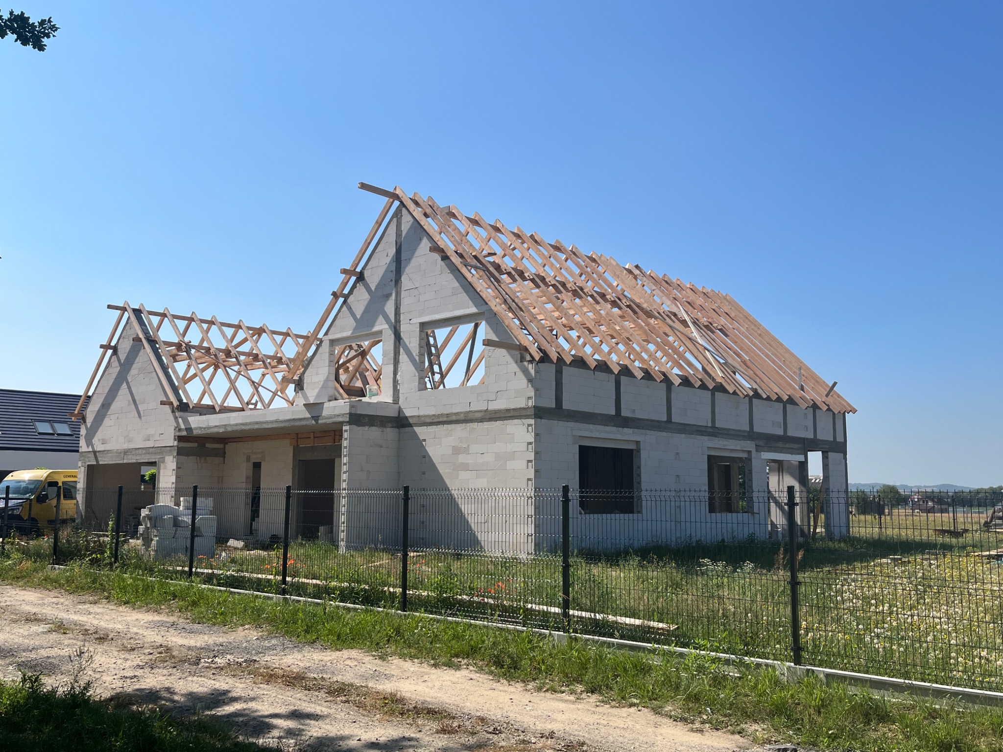 Surowy stan domu z białego bloczka z drewnianą konstrukcją dachu w trakcie budowy, widok z zewnątrz za metalowym ogrodzeniem, w tle błękitne niebo i żółty samochód dostawczy.
