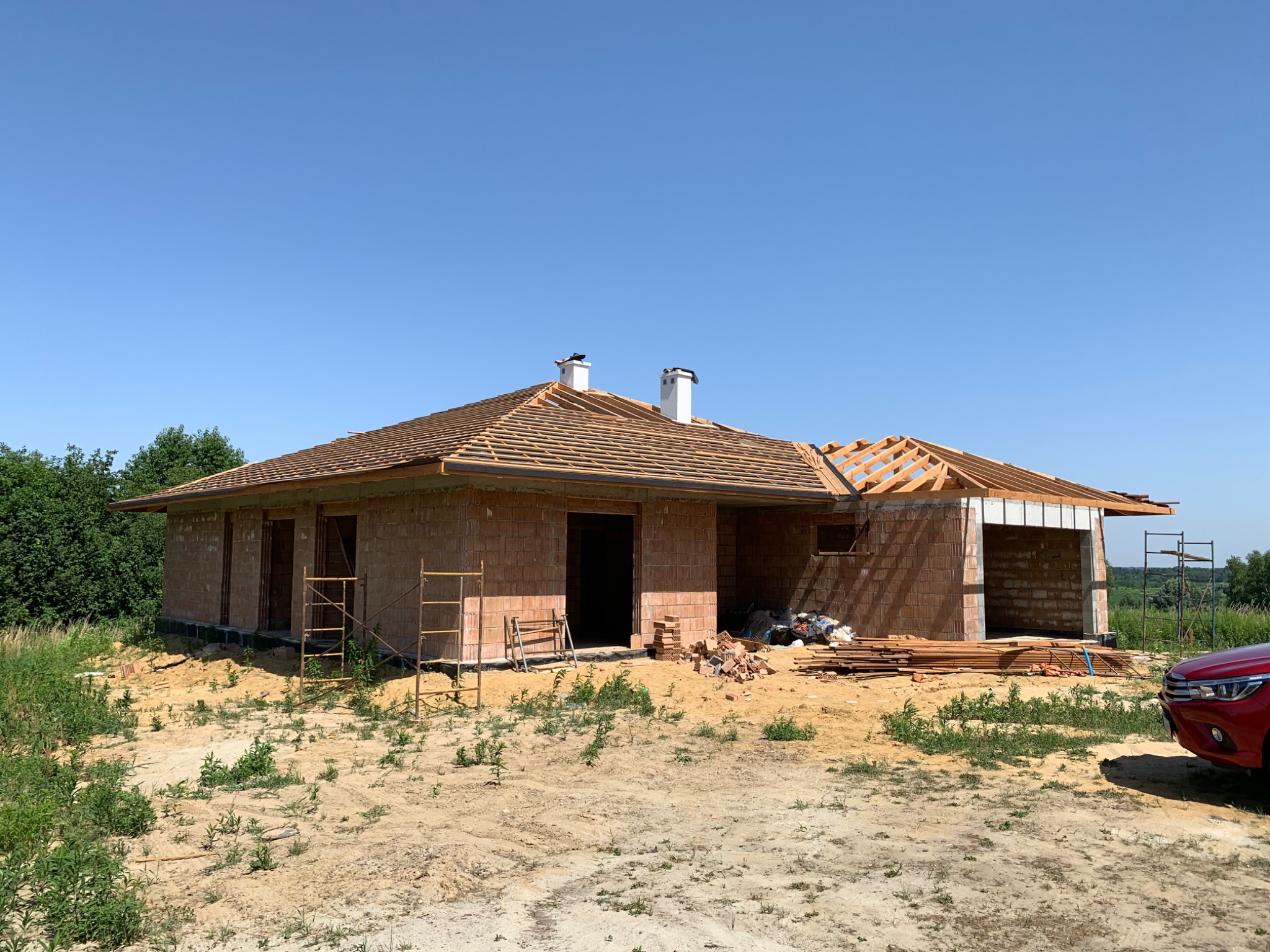 Budowa parterowego domu z czerwonej cegły, widoczne drewniane elementy konstrukcji dachu, białe kominy, teren budowy z rozrzuconymi materiałami.