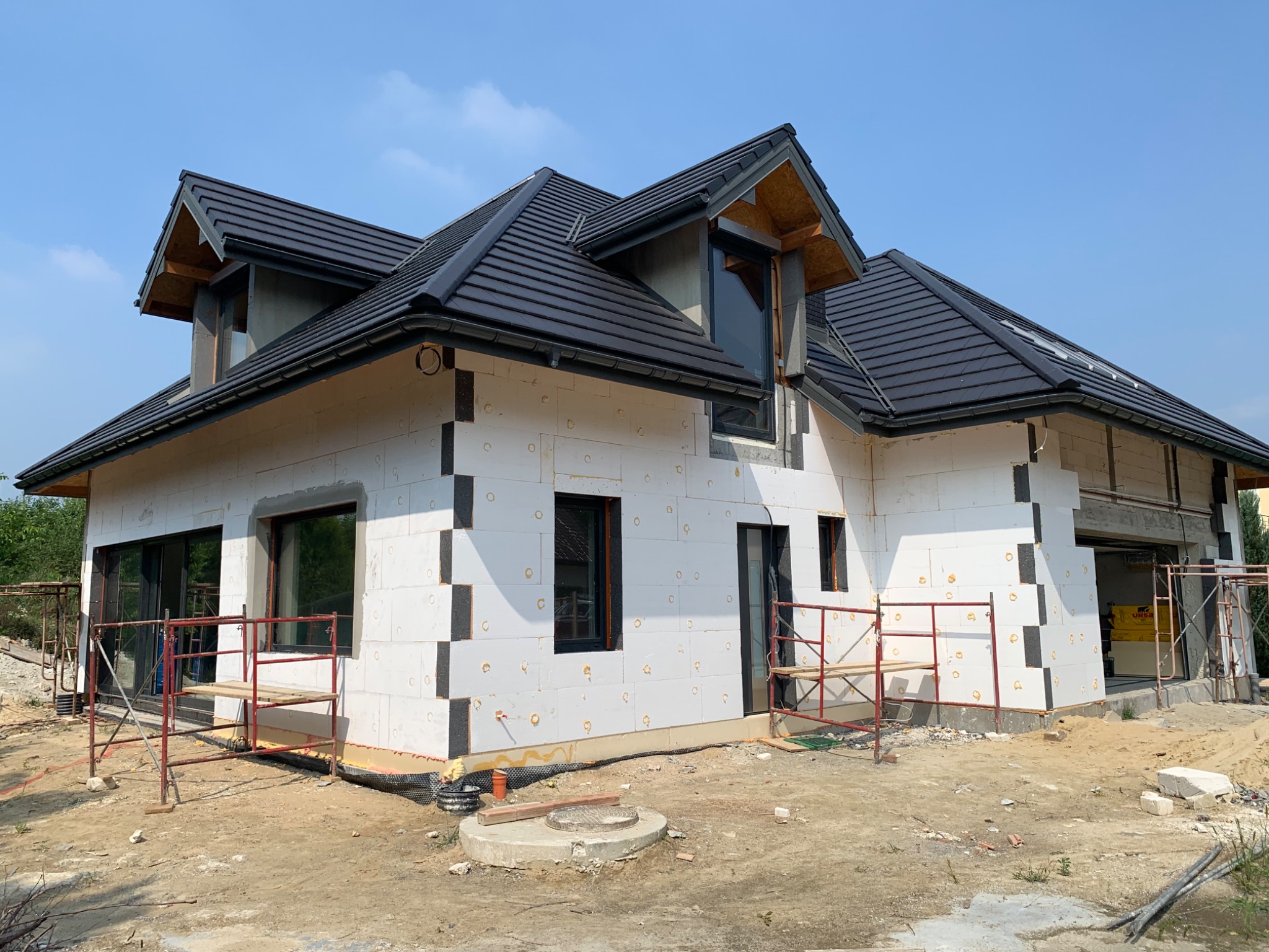 Niedokończony dom jednorodzinny z czarną dachówką, ocieplony białym styropianem z grafitowymi elementami, widoczne rusztowania i teren budowy.