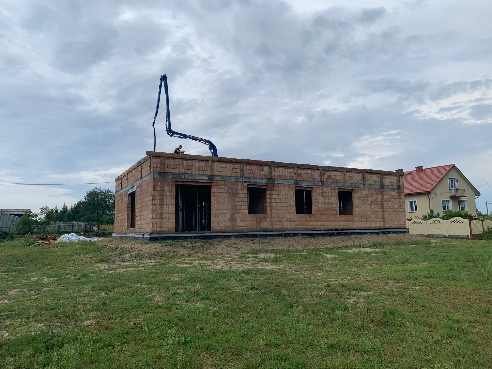 Wznoszony budynek z czerwonej cegły ceramicznej, widoczny operator pompy do betonu na dachu, w tle dom jednorodzinny z czerwonym dachem i szarym niebem.