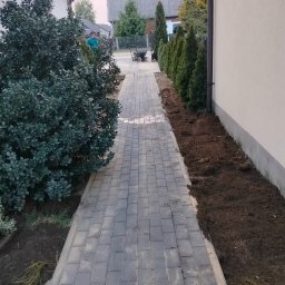 Marcin - Ułożona kostka brukowa tworząca ścieżkę w ogrodzie, z przygotowanym miejscem na nasadzenia po bokach. Widoczna osoba z wózkiem w tle.