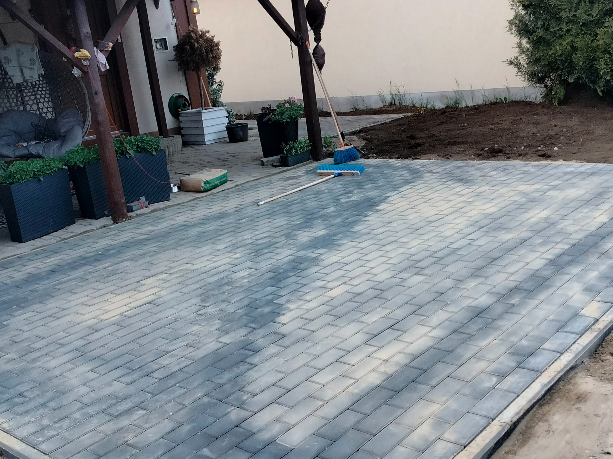 Świeżo ułożona, szara kostka brukowa na podwórku, widok z perspektywy. W tle widoczne rośliny w donicach, elementy drewnianej konstrukcji i narzędzia pracy.