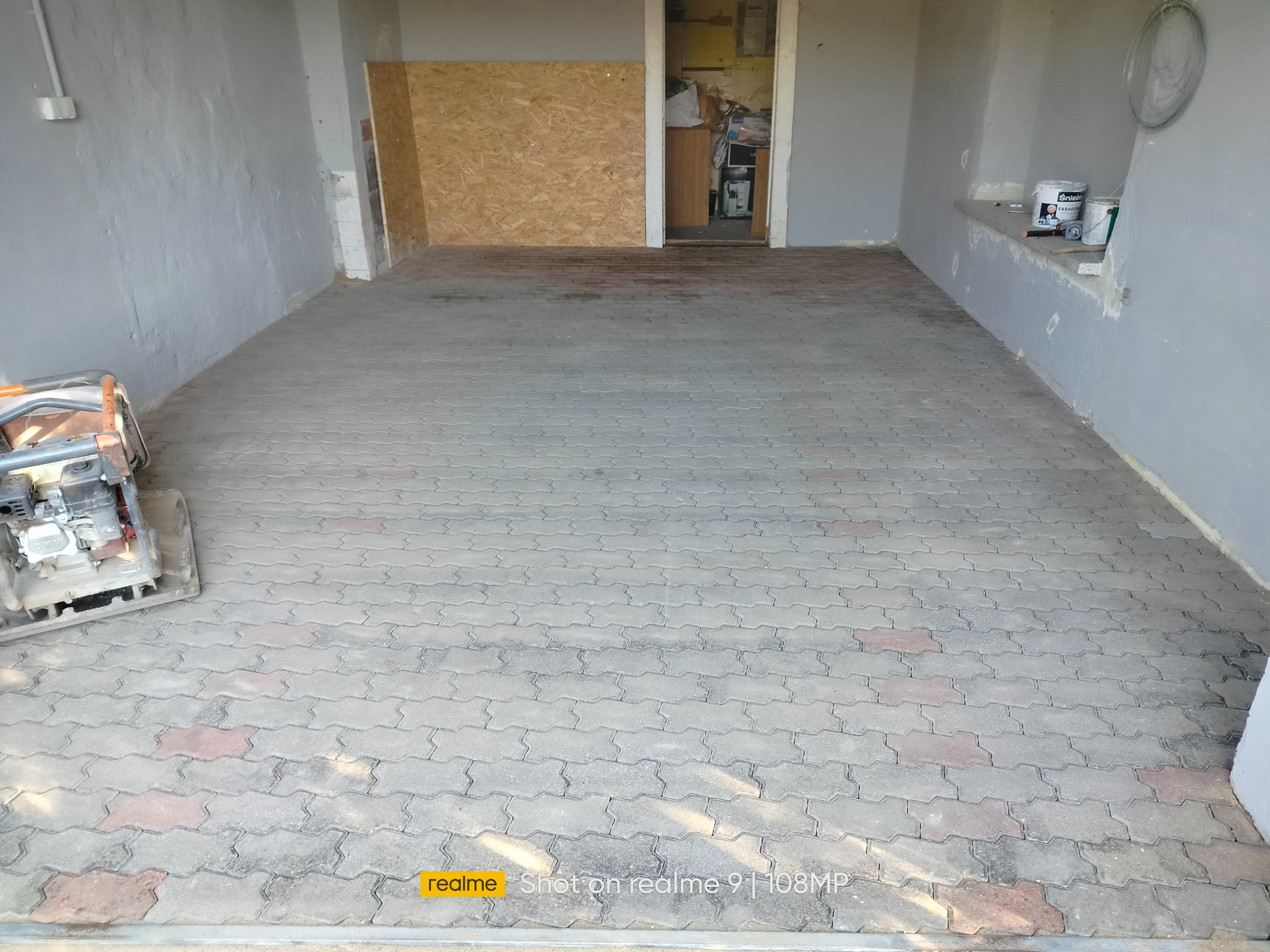 Ułożona kostka brukowa w garażu z szarą ścianą i widoczną ubijarką. Częściowo widoczne puszki z farbą. Kostka w odcieniach szarości z elementami czerwieni.