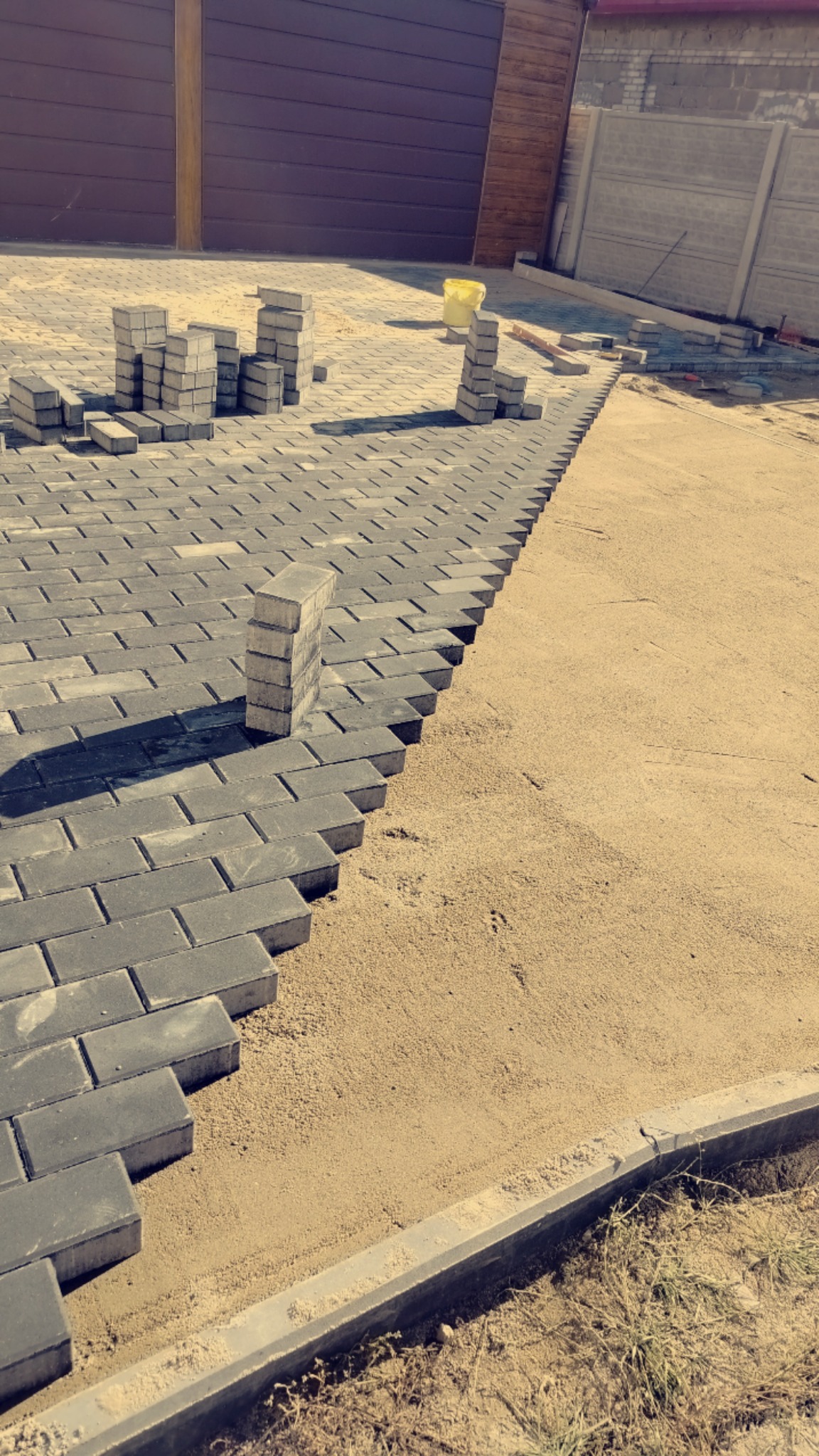 Układanie szarej kostki brukowej na piasku, widoczne stosy kostki i krawężnik. Częściowo ułożona nawierzchnia, etap prac. Tło: brama i ogrodzenie.