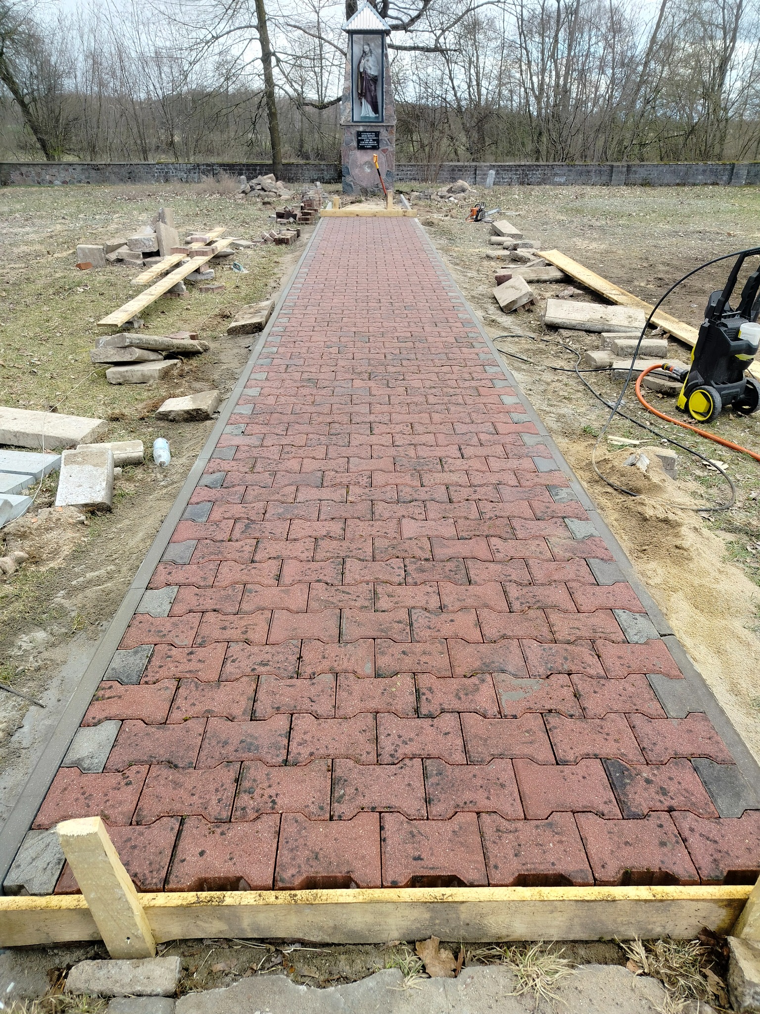 Czerwona kostka brukowa w jodełkę, tworząca ścieżkę w kierunku kapliczki. Widoczne drewniane szalunki i narzędzia. Częściowo ukończona realizacja.