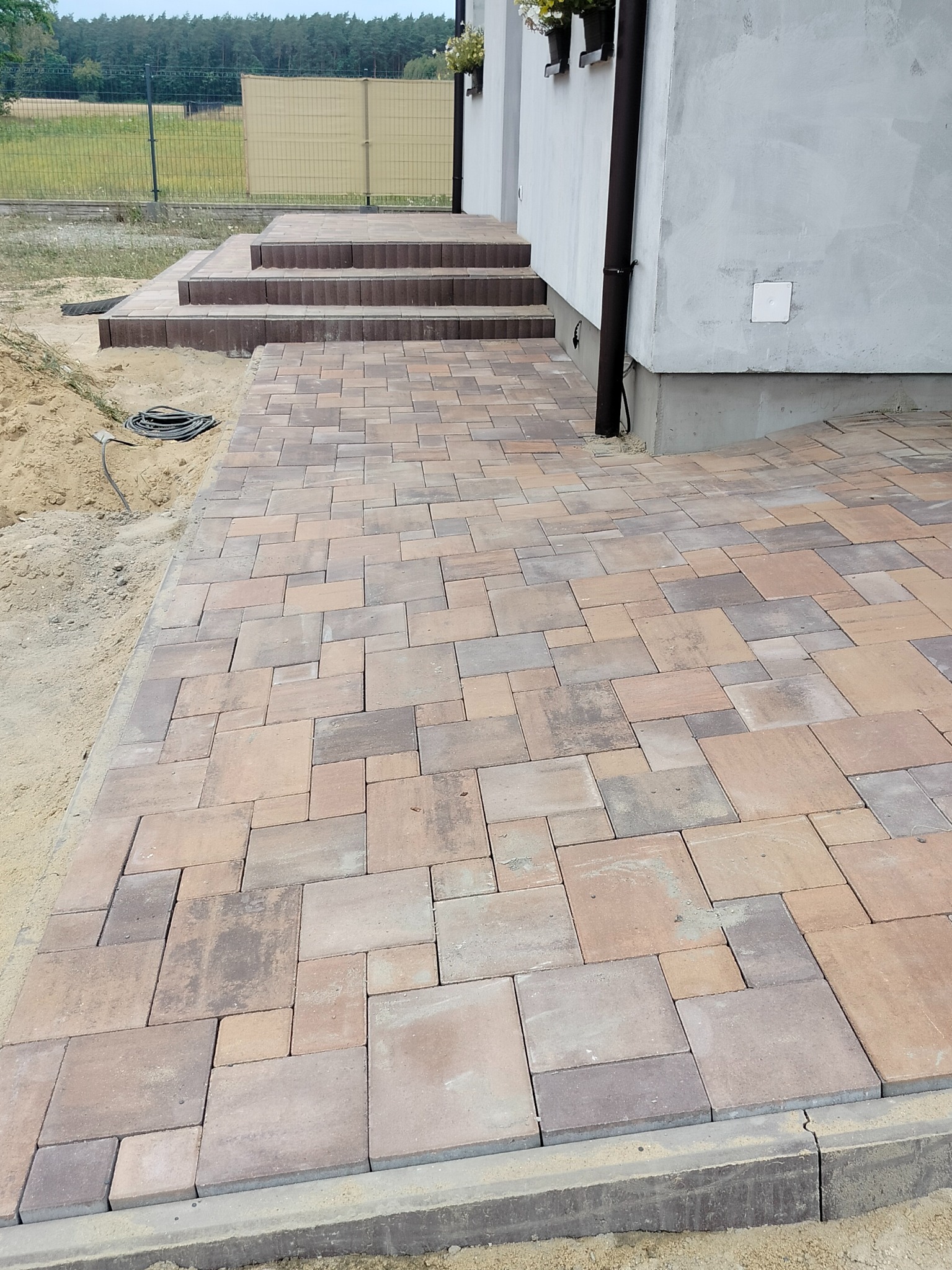 Ułożona kostka brukowa w odcieniach brązu i beżu prowadzi do schodów wejściowych domu. Widoczny fragment elewacji i trawnik w tle. Estetyczne wykończenie.