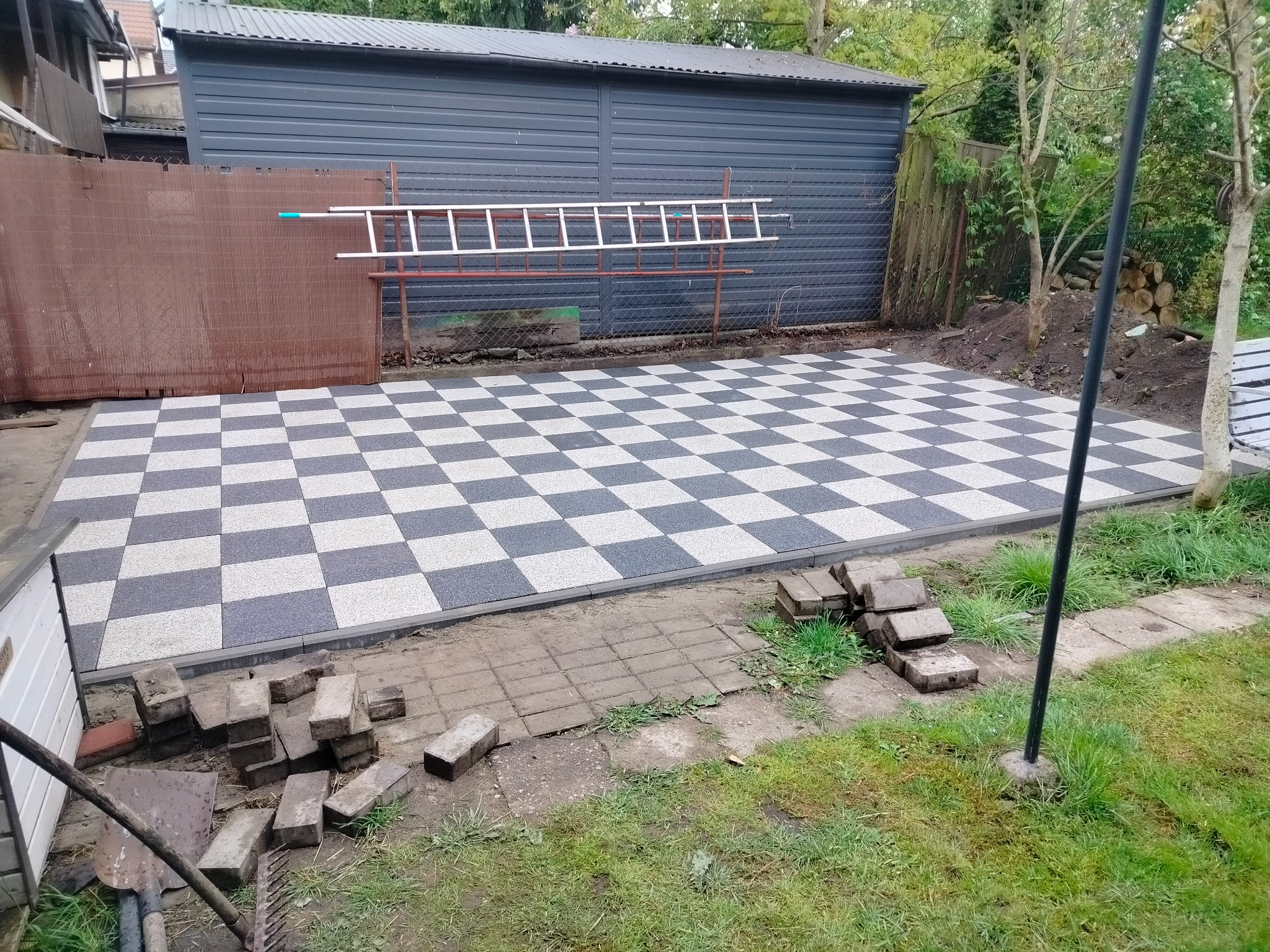 Szachownica z kostki brukowej w ogrodzie, z drabiną opartą o blaszany garaż. Część kostki jeszcze nieułożona, widoczne narzędzia i trawa.