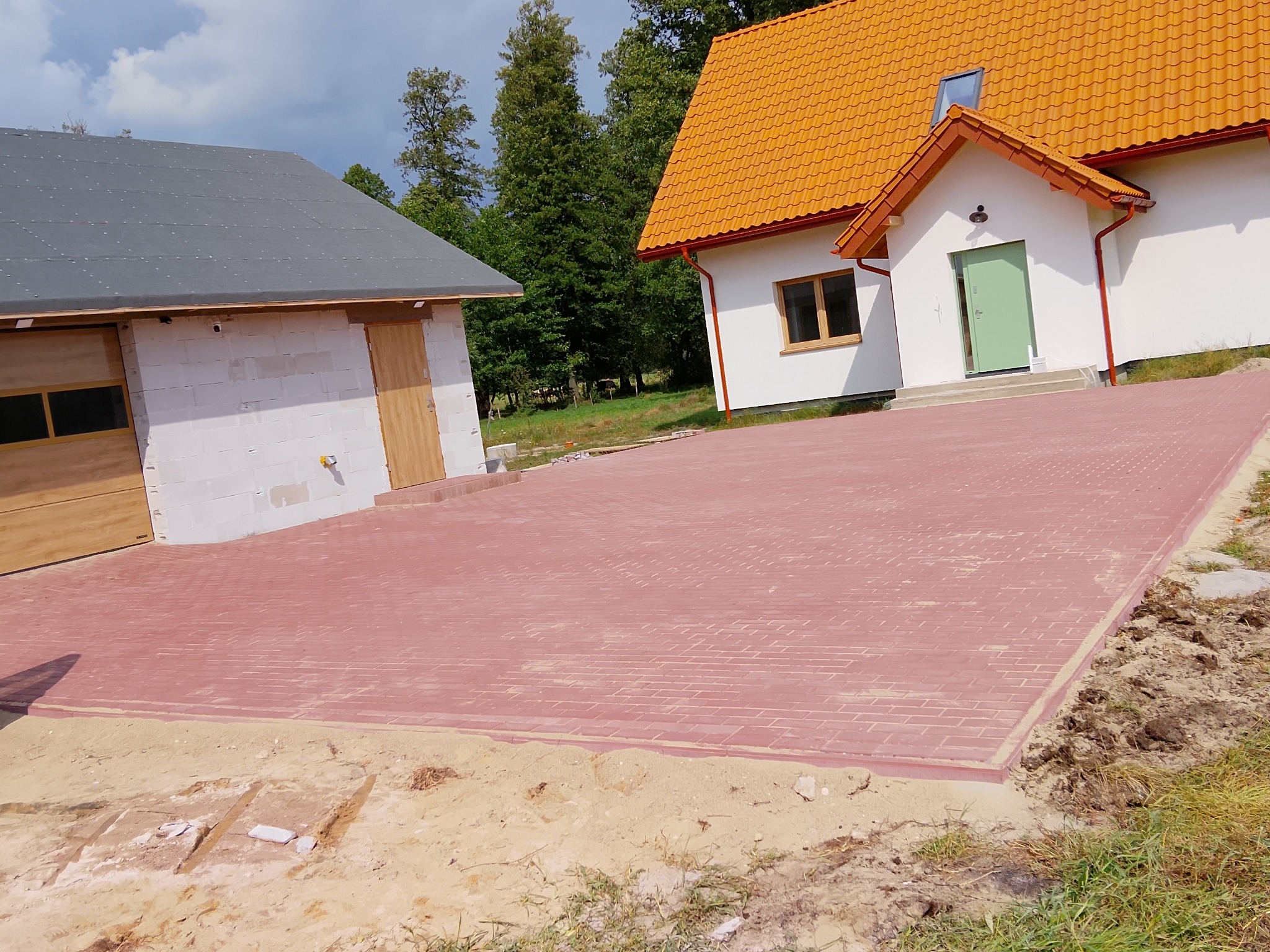 Czerwona kostka brukowa ułożona na podjeździe przed nowym domem z pomarańczowym dachem i garażem z białego bloczka. Widoczne fragmenty trawnika i piasku.