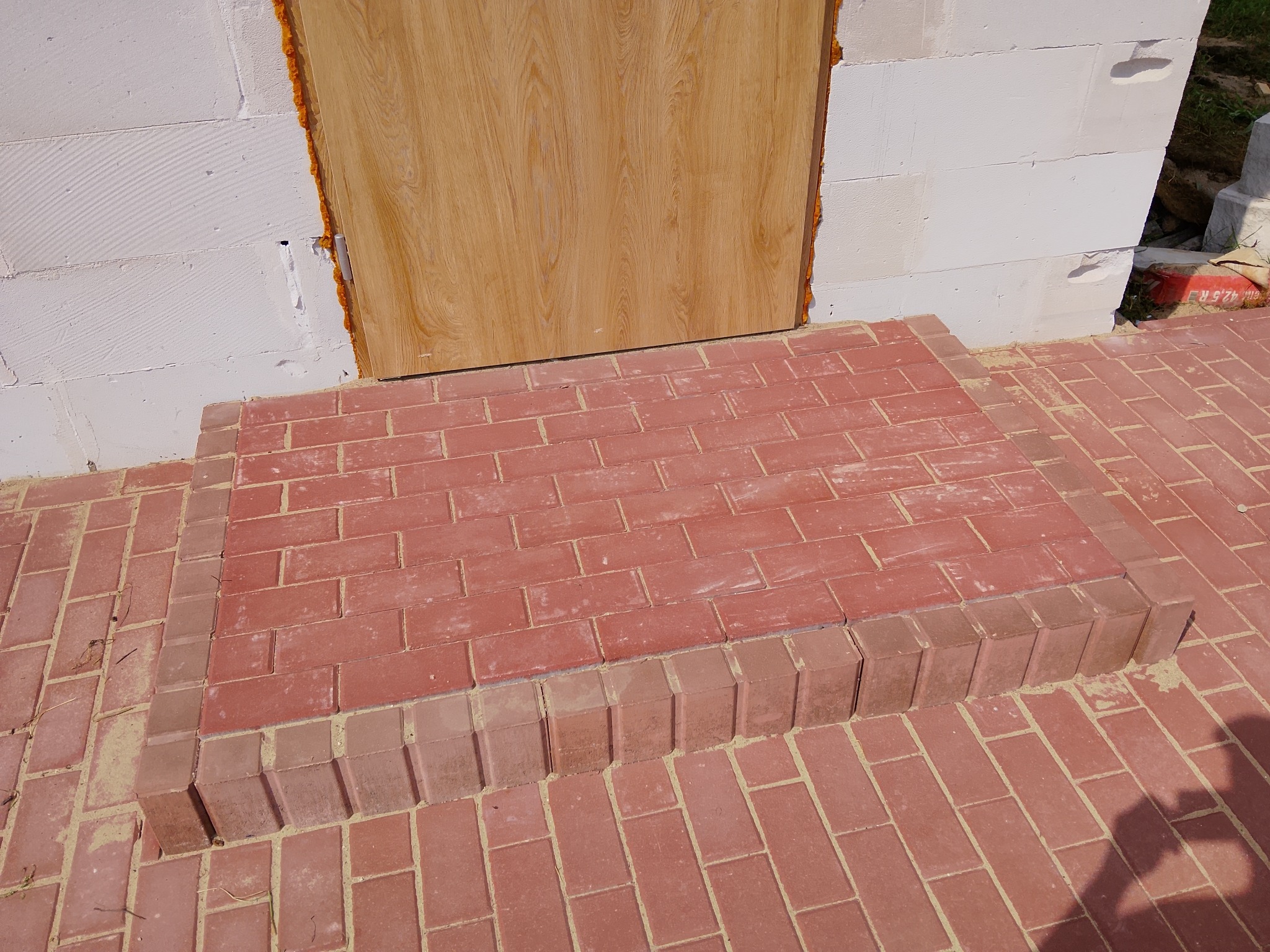 Czerwona kostka brukowa tworzy schody wejściowe do budynku z białych bloczków. Widoczna pianka montażowa wokół ościeżnicy. Detal wykończenia.