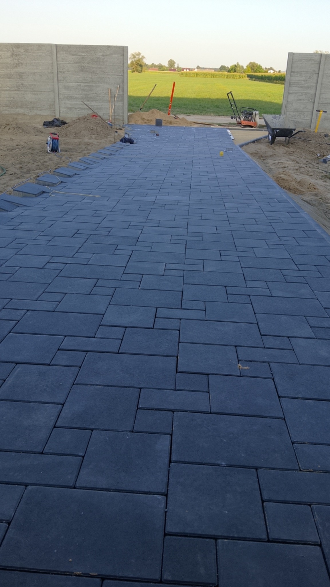 Ciemna kostka brukowa ułożona na ścieżce, widok z perspektywy. W tle brama z betonowych płyt i krajobraz polny. Częściowo ukończone prace brukarskie.