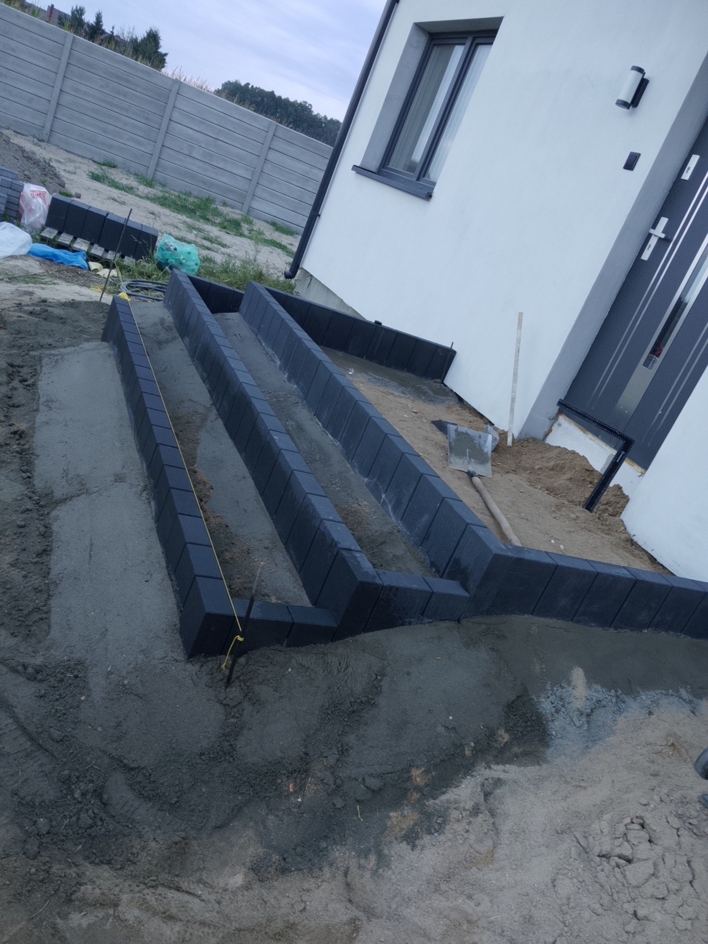 Wykonywanie obrzeży z kostki przy domu. Widoczne czarne kostki obrzeżowe, zaprawa, piasek, szpadel i fragment ogrodzenia panelowego.