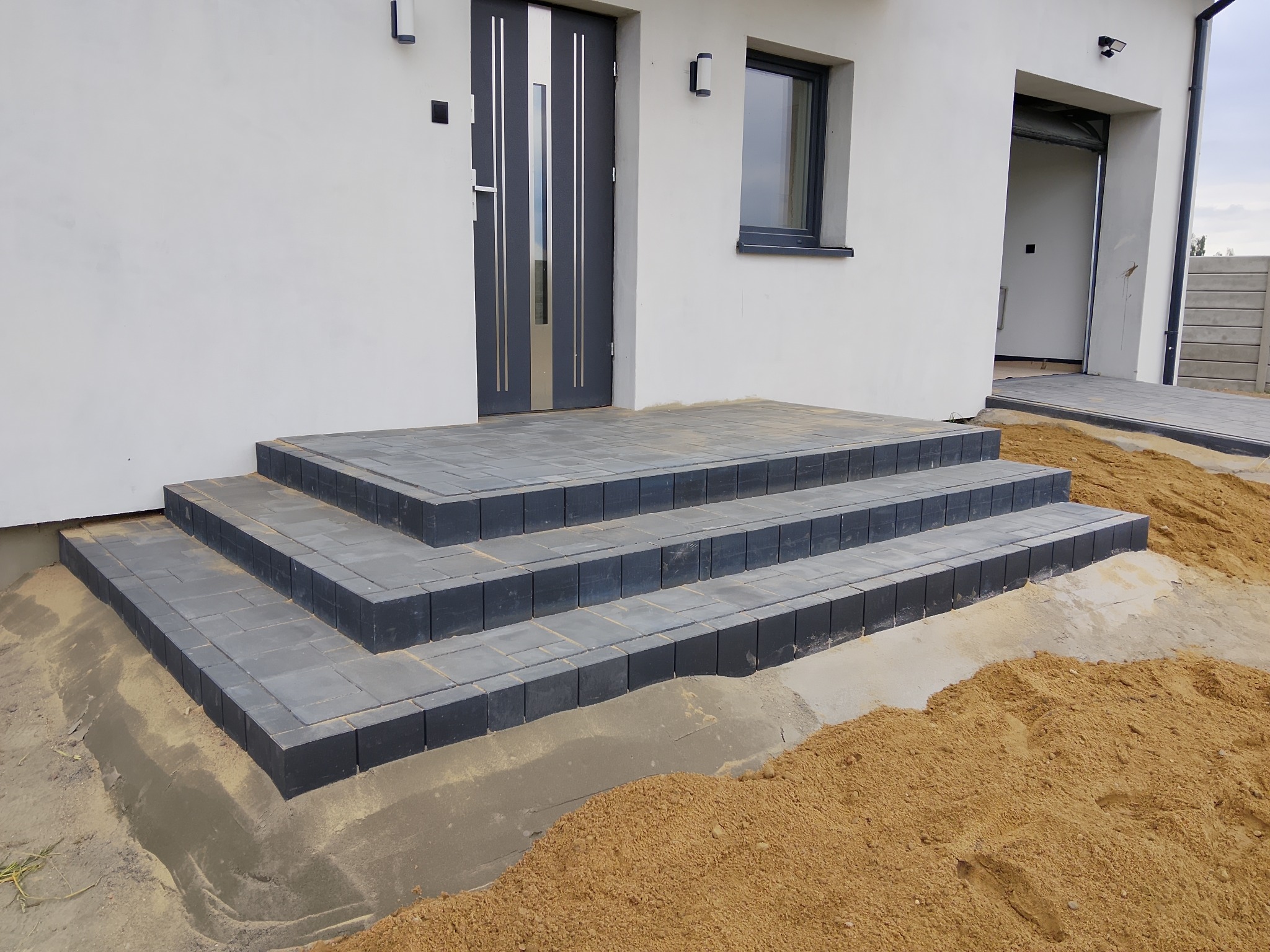 Nowoczesne schody wejściowe z ciemnej kostki brukowej, prowadzące do białego domu z szarą stolarką. Piasek budowlany wokół, częściowo ułożona kostka przed garażem.