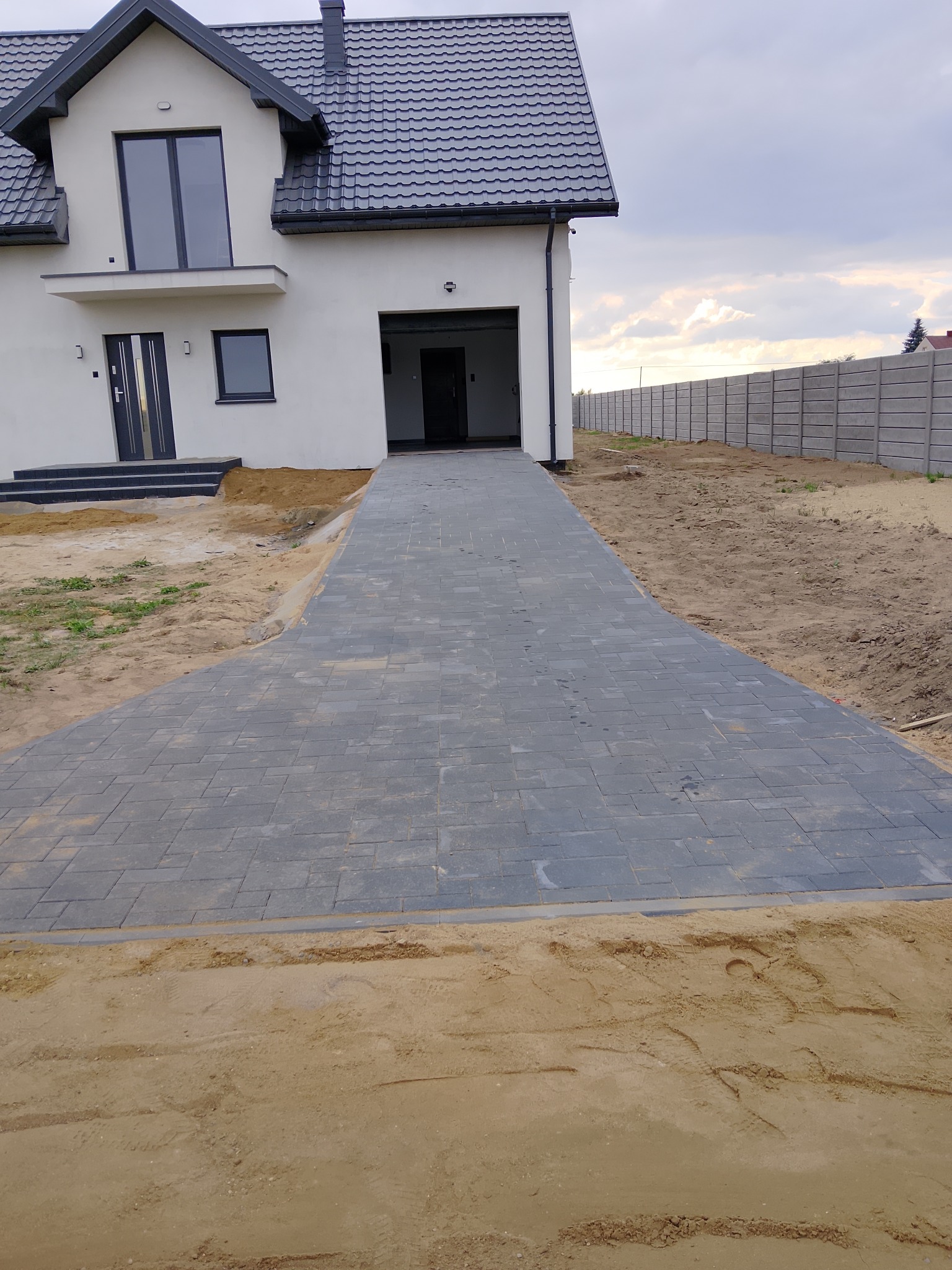 Nowoczesny dom z szarą kostką brukową na podjeździe i betonowym ogrodzeniem w tle, widok z poziomu gruntu.