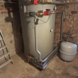 Szary akumulator ciepła z rurami i pompami w piwnicy z ceglaną ścianą, obok drabina i zbiornik wyrównawczy.