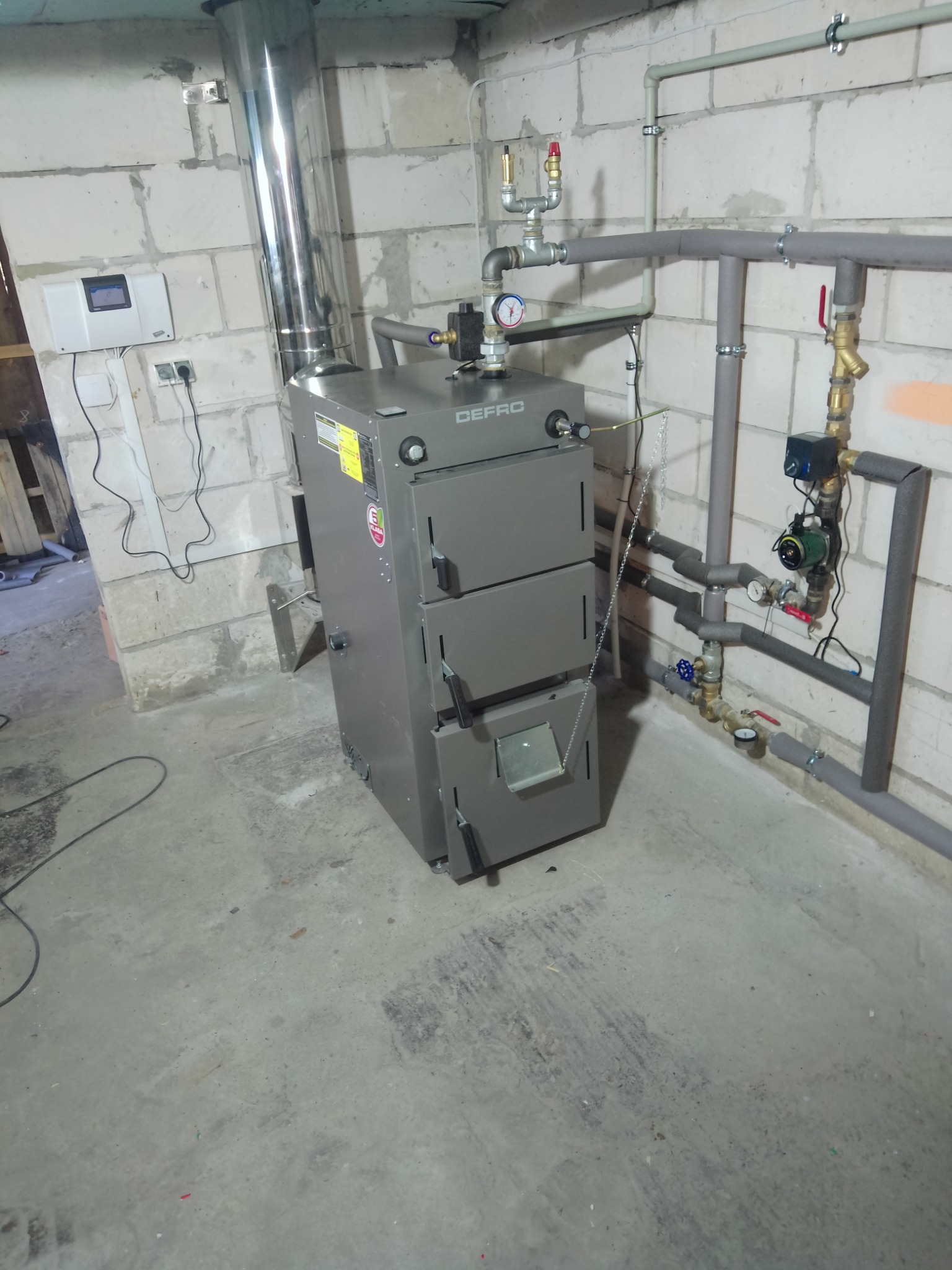 Szary piec CO marki Defro z podłączonym kominem i instalacją grzewczą w surowym pomieszczeniu z betonową podłogą i ścianami z bloczków.