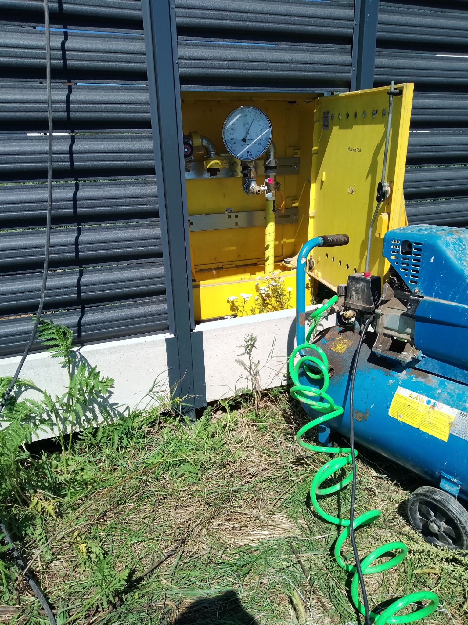 Żółta szafka gazowa z otwartymi drzwiczkami, widoczny manometr i zawory, obok niebieski kompresor z zielonym wężem spiralnym na trawie.