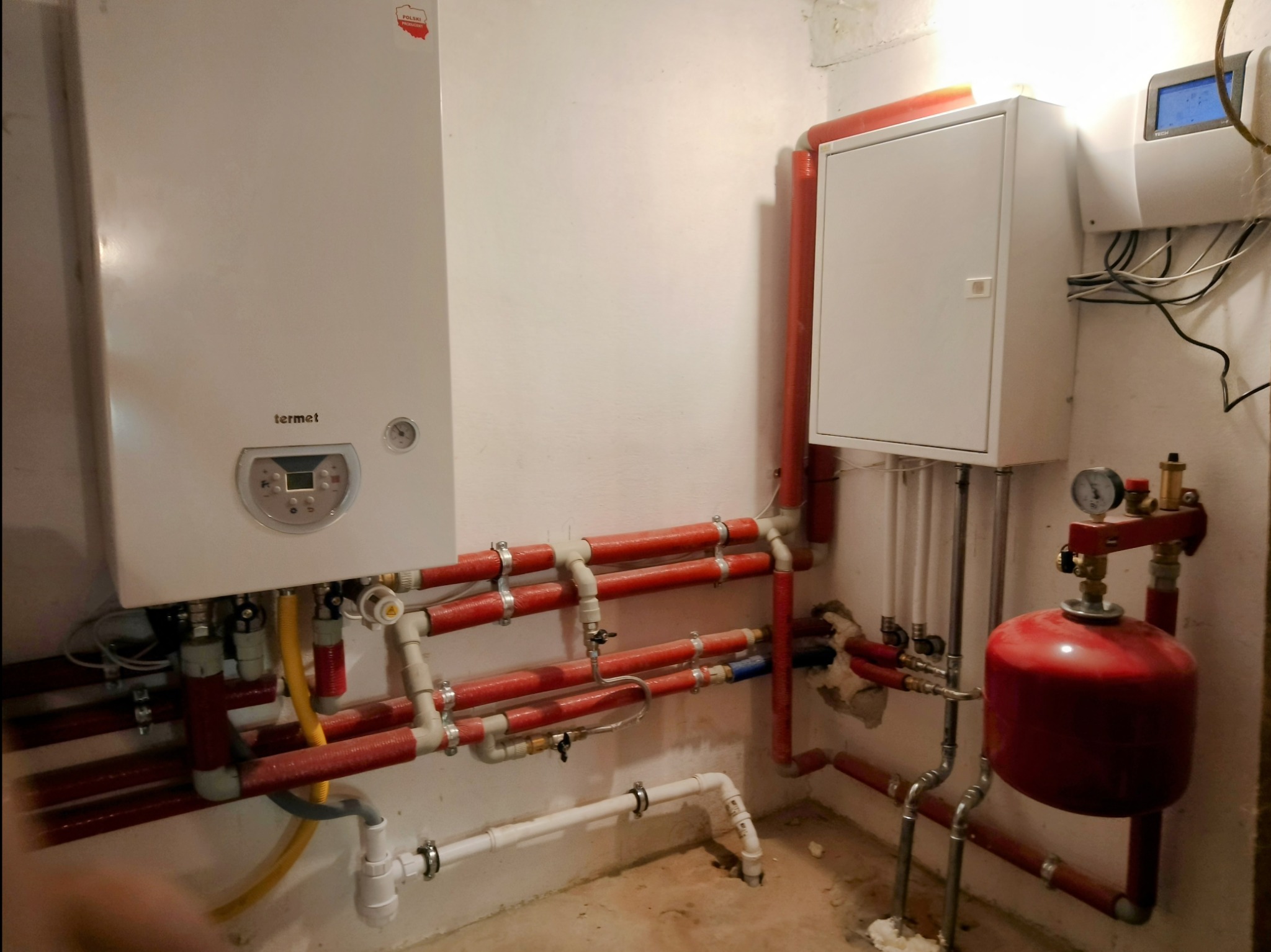 Biały piec gazowy Termet z podłączonymi rurami grzewczymi w kolorze czerwonym, widoczny również biały zbiornik wyrównawczy i skrzynka sterownicza na ścianie w pomieszczeniu technicznym.