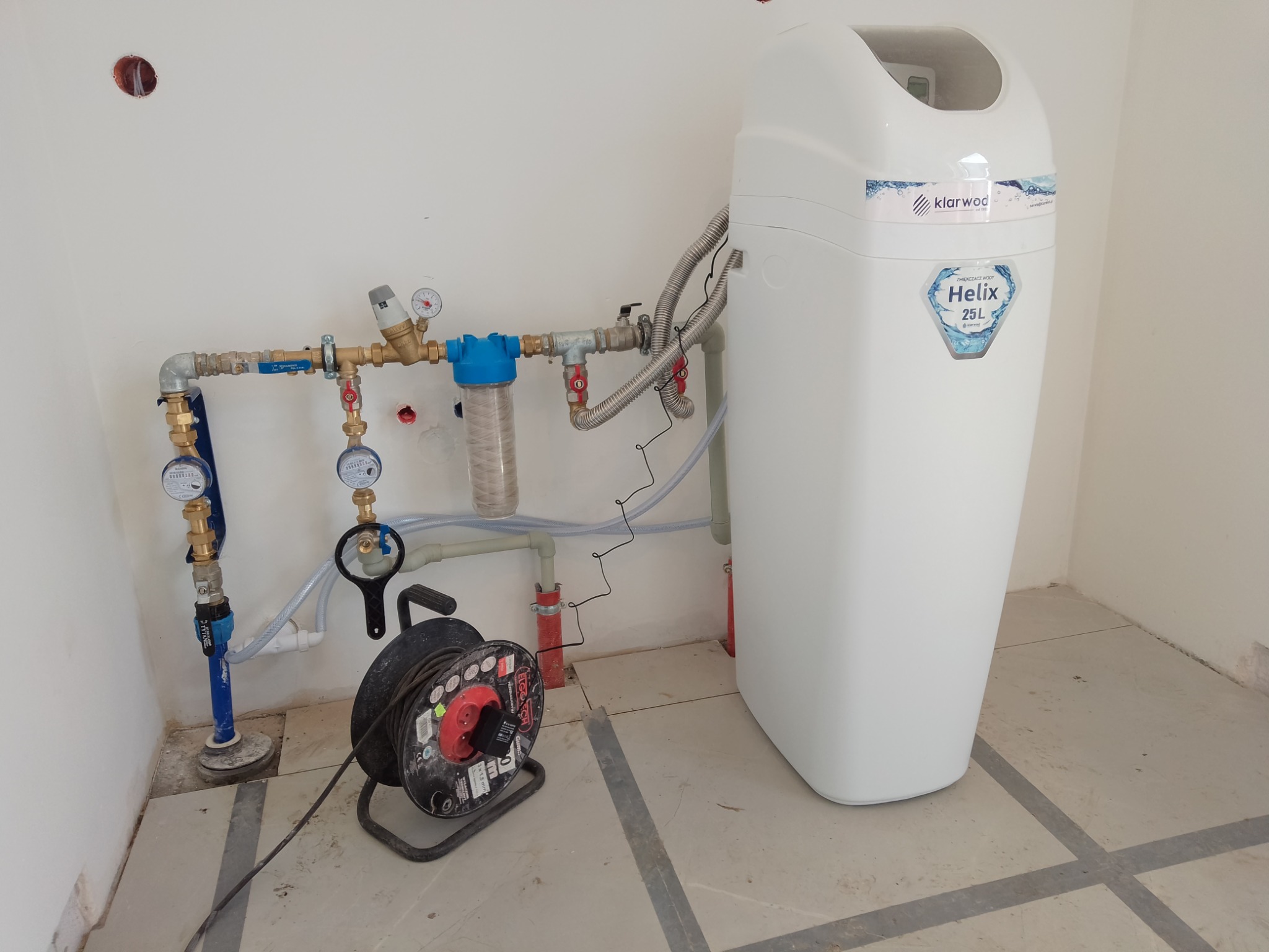 Biały zmiękczacz wody Klarwod Helix 25L podłączony do instalacji wodnej z filtrem i manometrami, z widoczną zwiniętą przedłużką elektryczną.