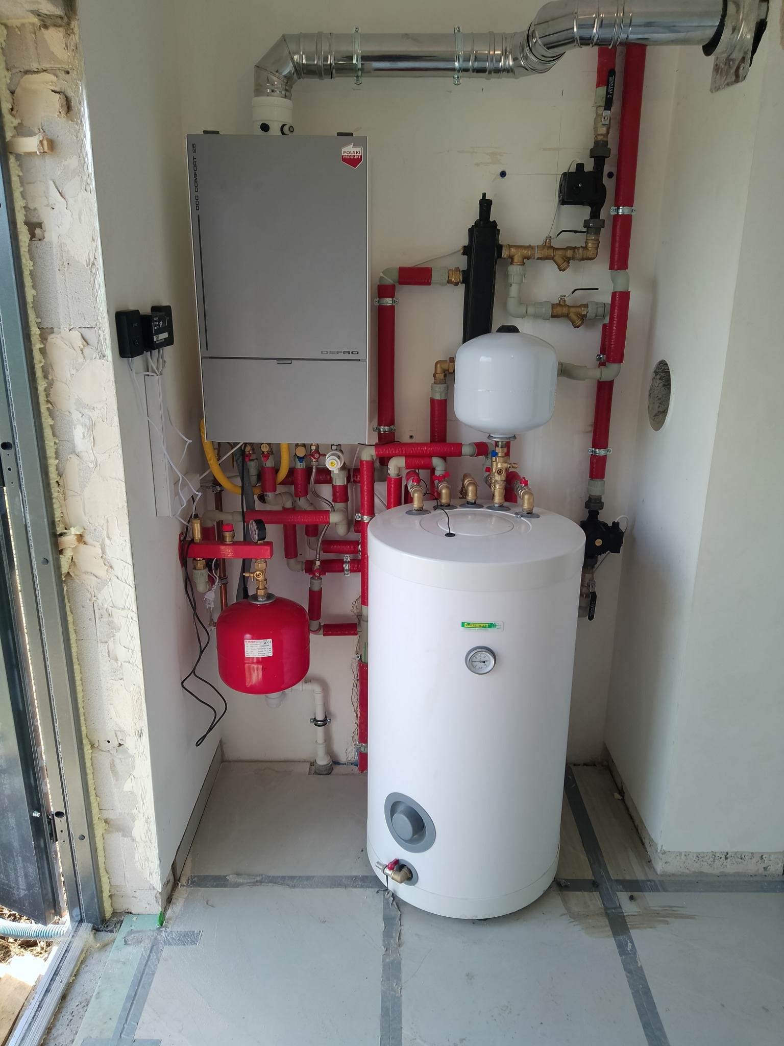 Nowoczesny piec gazowy Defro z zasobnikiem ciepłej wody, instalacją z czerwonych rur i osprzętem, zamontowany w nowym pomieszczeniu w trakcie wykańczania.