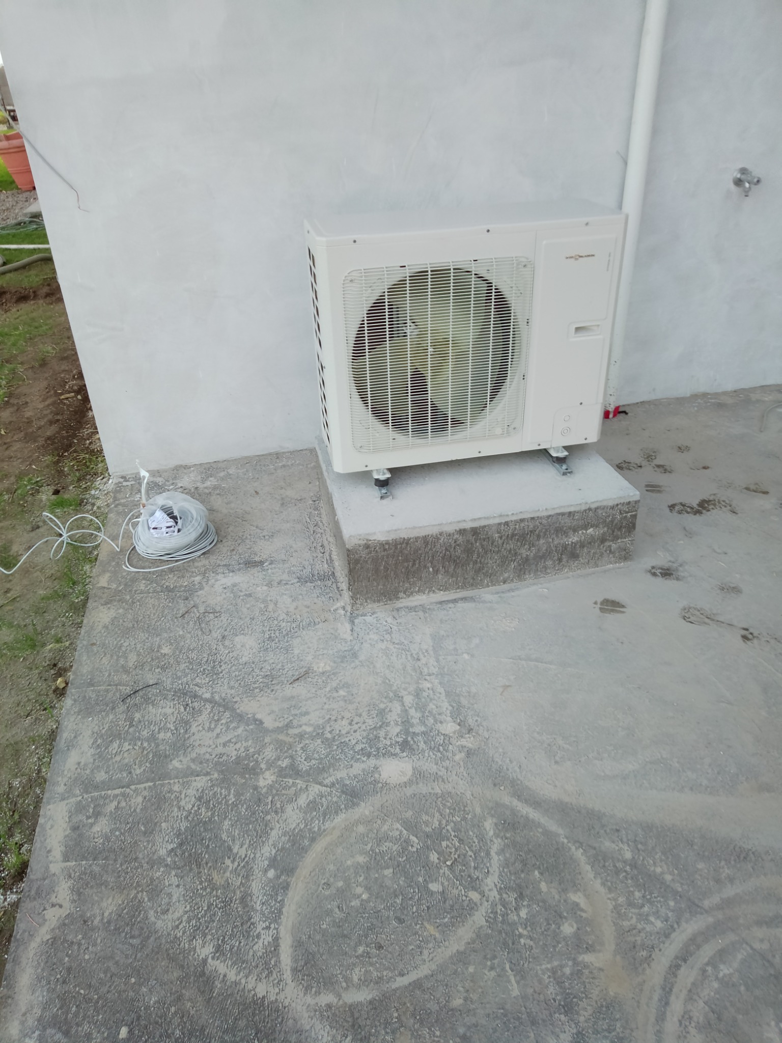 Zewnętrzna jednostka klimatyzacji Viessmann zamontowana na betonowym cokole przy szarej ścianie budynku, obok zwój kabla elektrycznego.