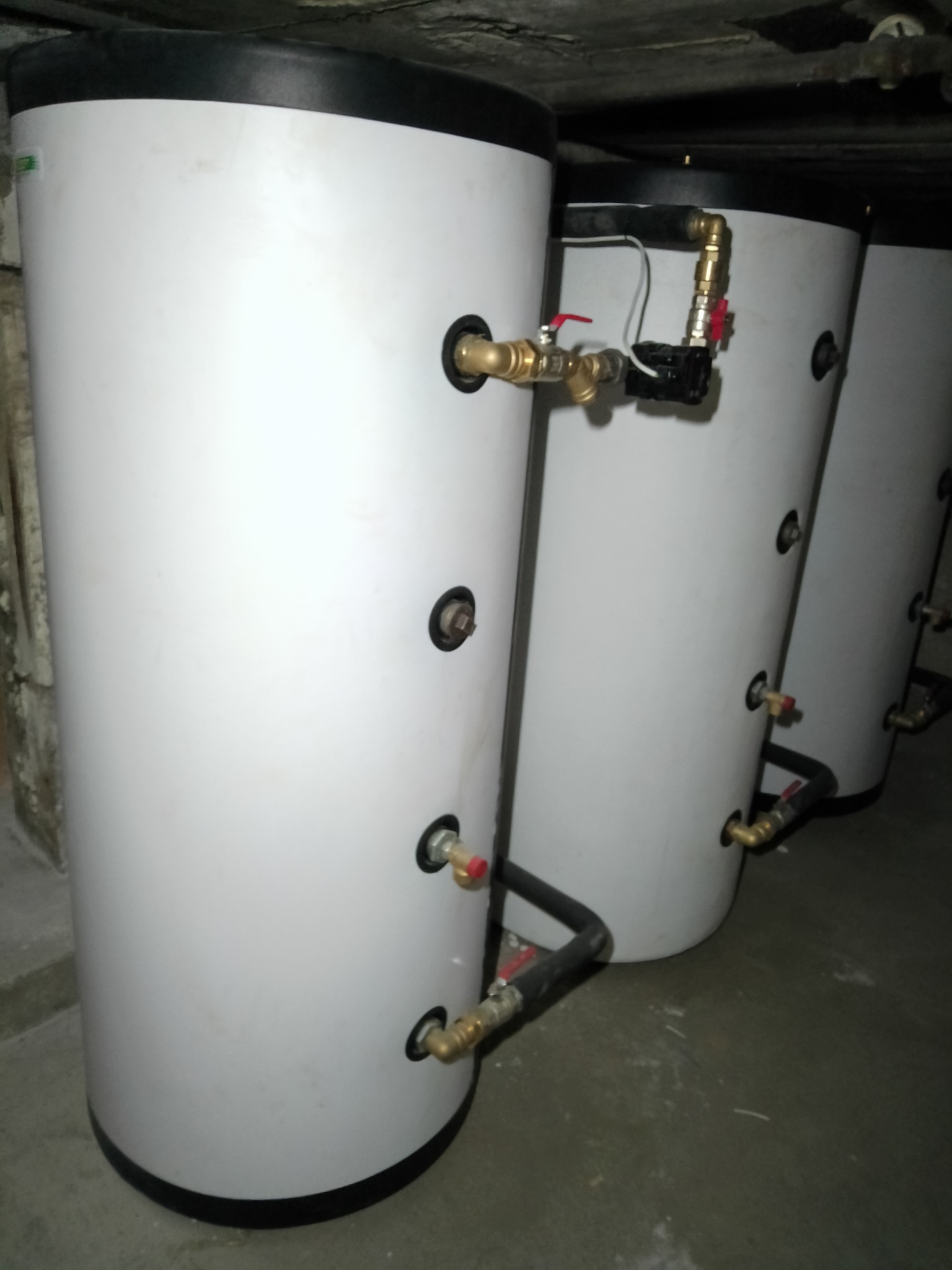 Białe, cylindryczne zbiorniki akumulacyjne z czarnymi pokrywami i podłączeniami hydraulicznymi, ustawione w rzędzie w piwnicy o surowym wykończeniu.