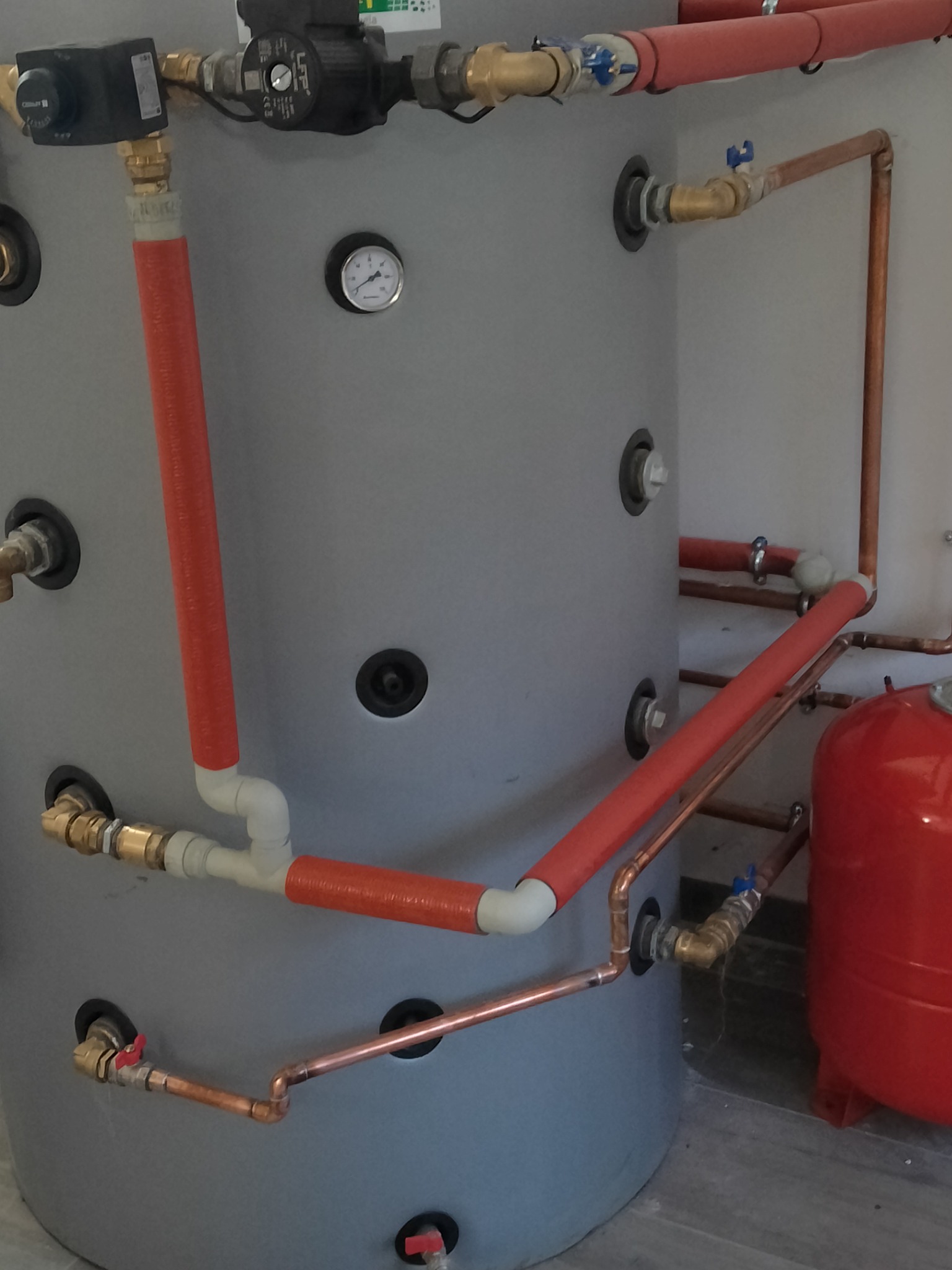 Szary zbiornik akumulacyjny z podłączonymi rurami miedzianymi i plastikowymi w izolacji termicznej, widoczny manometr i pompa obiegowa.