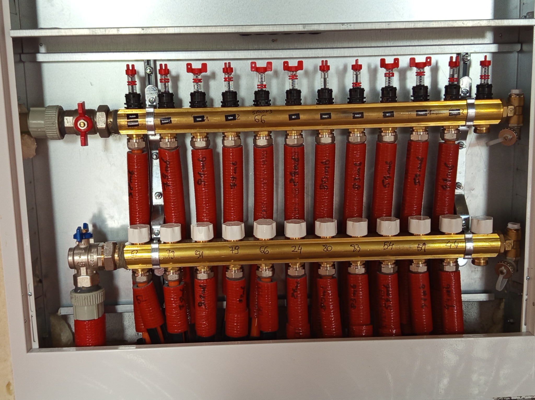 Rozdzielacz ogrzewania podłogowego w szafce, z czerwonymi rurami oznaczonymi numerami pomieszczeń, zawory regulacyjne i złote kolektory, całość w szarej obudowie.