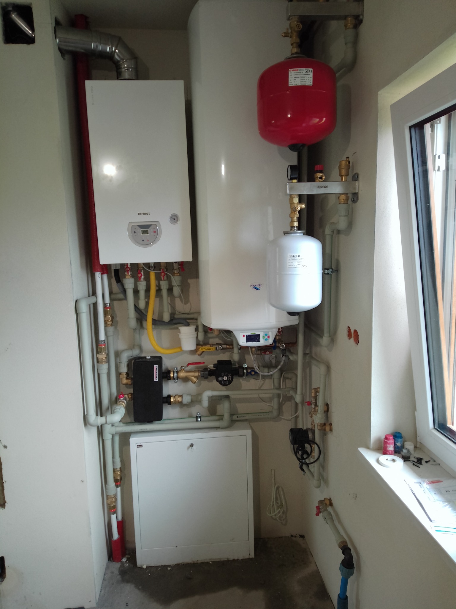 Złożona instalacja grzewcza z piecem gazowym Termet, zbiornikami i licznymi rurami, widoczna w pomieszczeniu z oknem. Elementy instalacji są nowe i czyste.