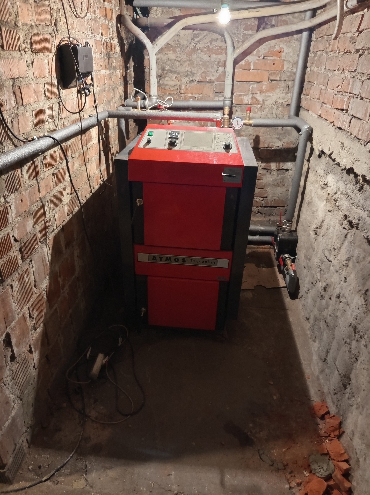 Czerwono-czarny piec Atmos z podłączonymi rurami w piwnicy z ceglanymi ścianami. Widoczne elementy instalacji i przewody elektryczne. Oświetlenie żarówką.