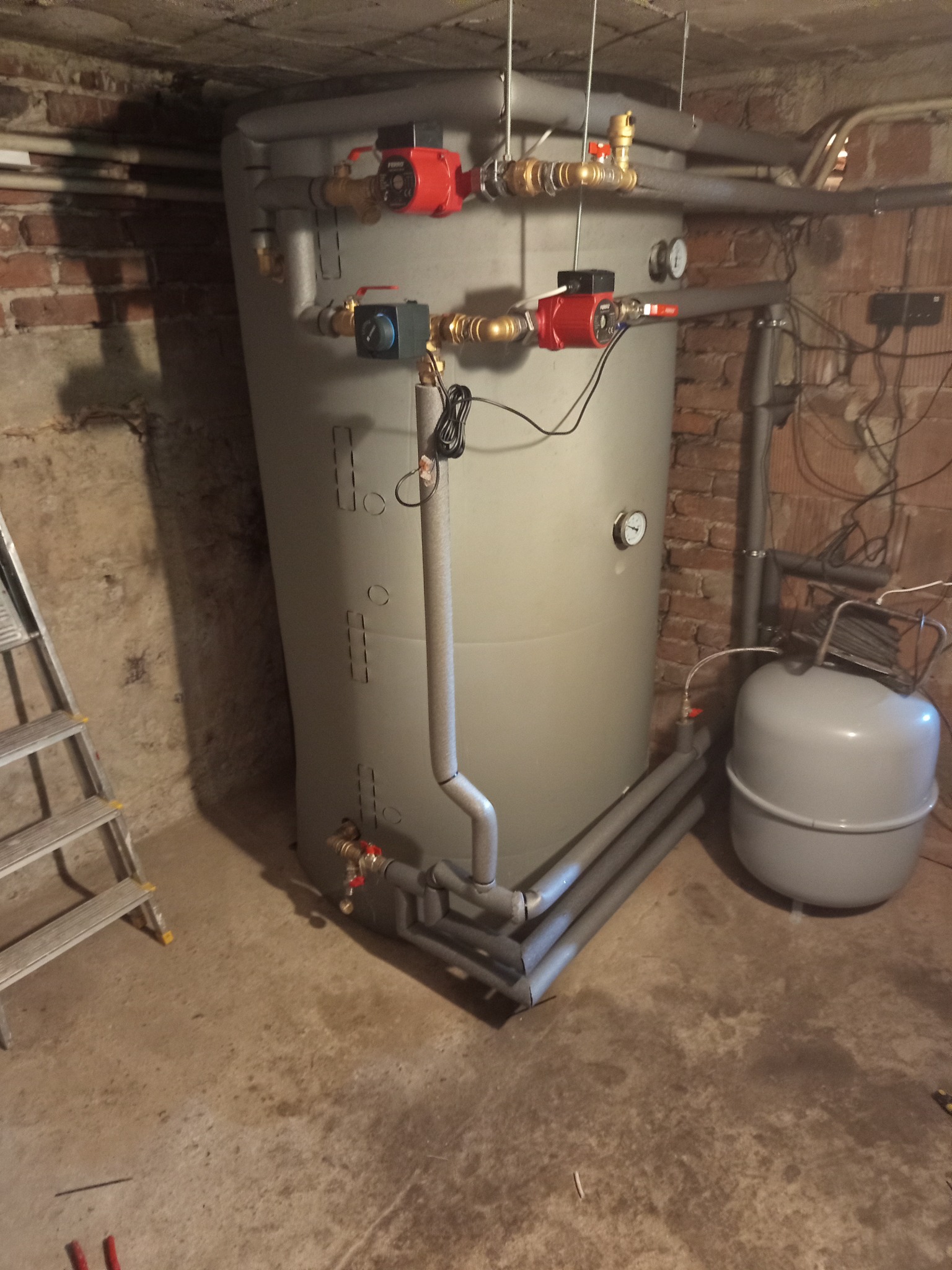 Szary akumulator ciepła z rurami i pompami w piwnicy z ceglaną ścianą, obok drabina i zbiornik wyrównawczy.
