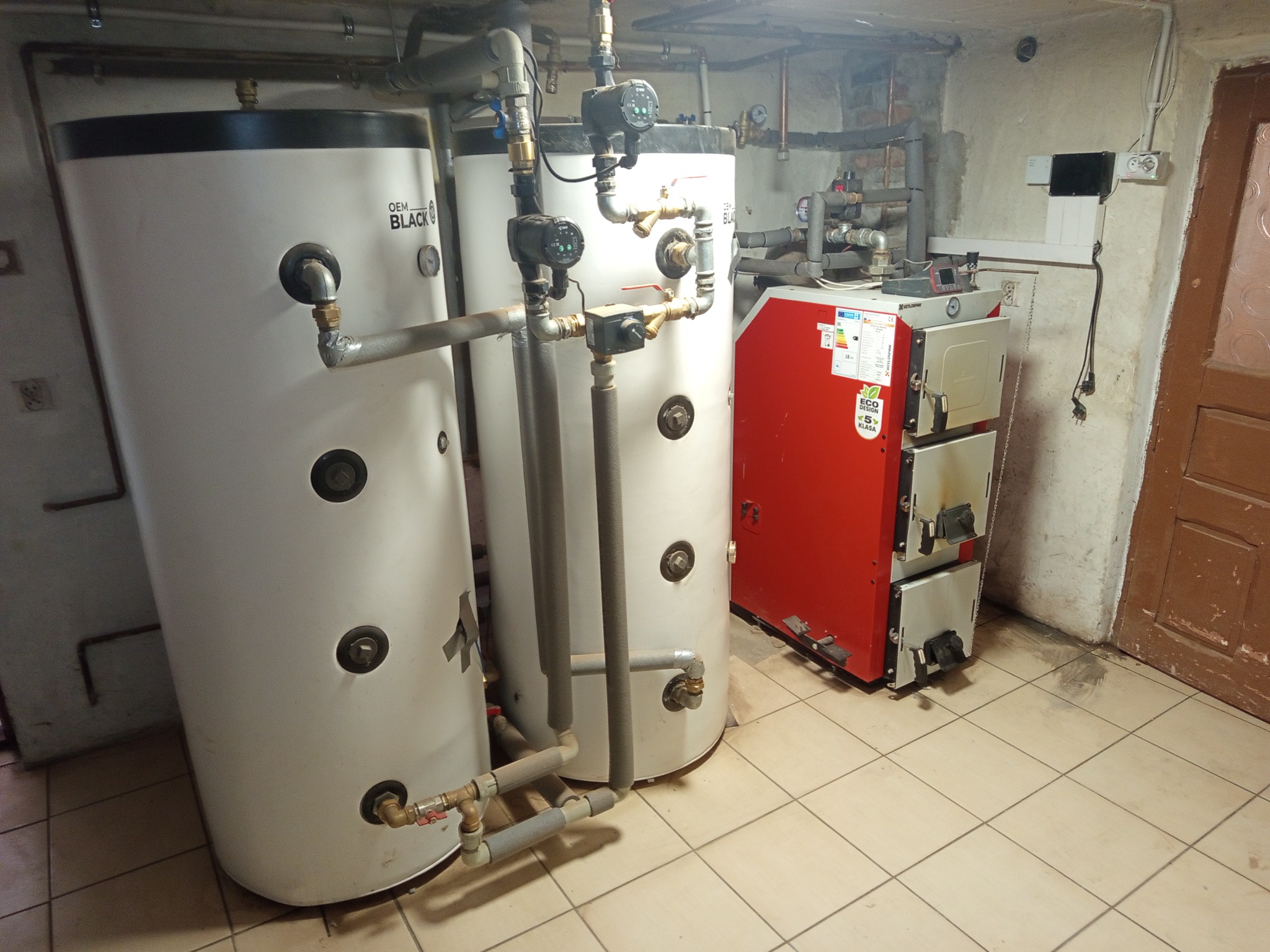 Dwa białe zbiorniki akumulacyjne OEM Black z rurami i zaworami, obok czerwony piec CO, na tle beżowych płytek podłogowych i fragmentu ściany.